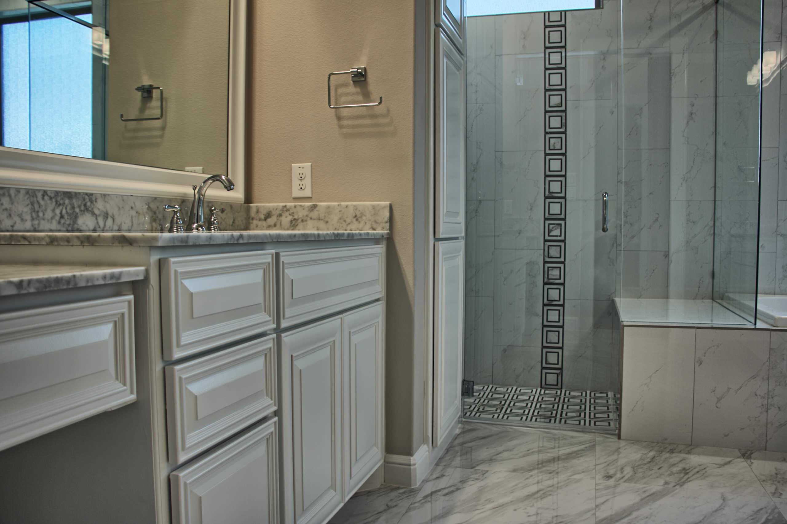White raised-panel vanity with marble top and chrome faucet beside a frameless glass shower with marble-look tile and black-and-white mosaic accents