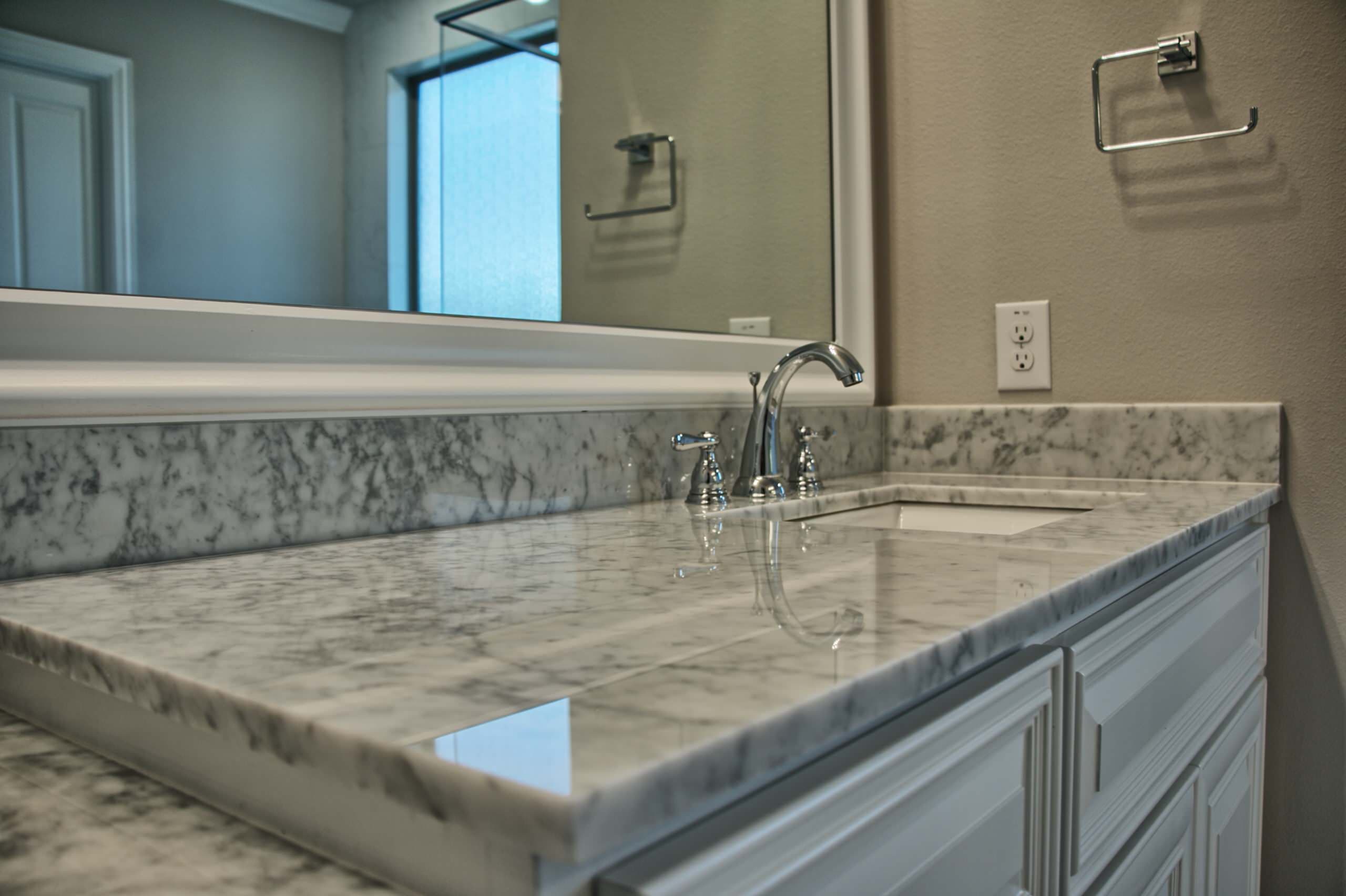 Close view of a white marble vanity top with undermount sink, chrome widespread faucet, white raised-panel cabinets and framed mirror