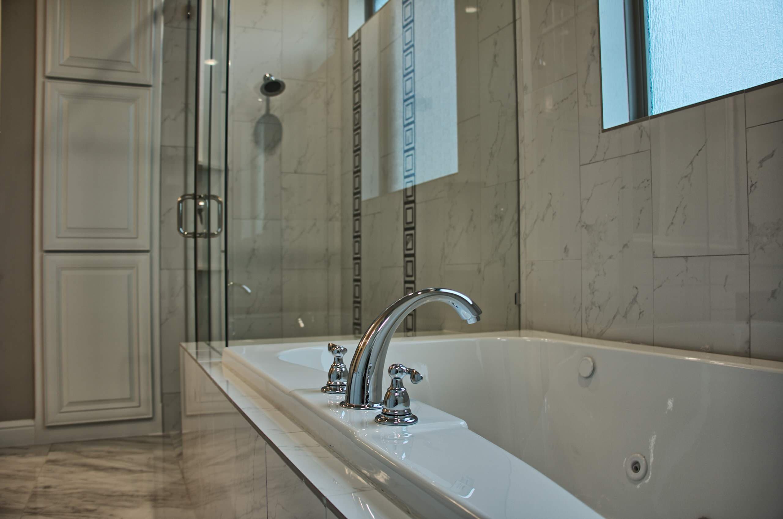 Chrome Roman faucet on a white jetted drop-in tub with marble-look tile surround, beside a glass shower with black-and-white mosaic accents