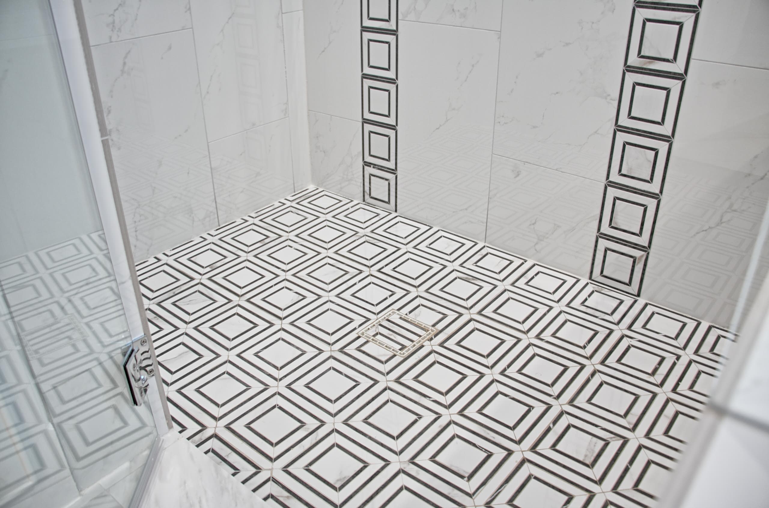 Shower floor in black-and-white geometric mosaic tile with square drain, framed by marble-look wall tile with black-and-white mosaic accent strips