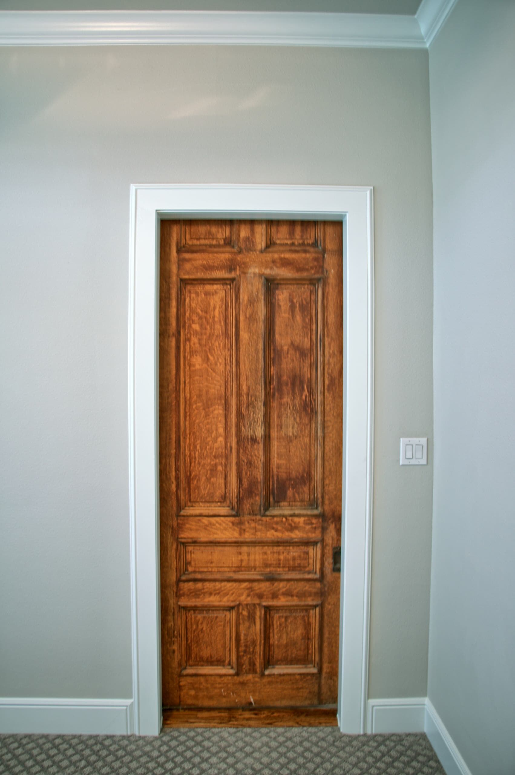 Stained oak paneled door in white casing set into a light gray wall, with crown molding above and patterned carpet below