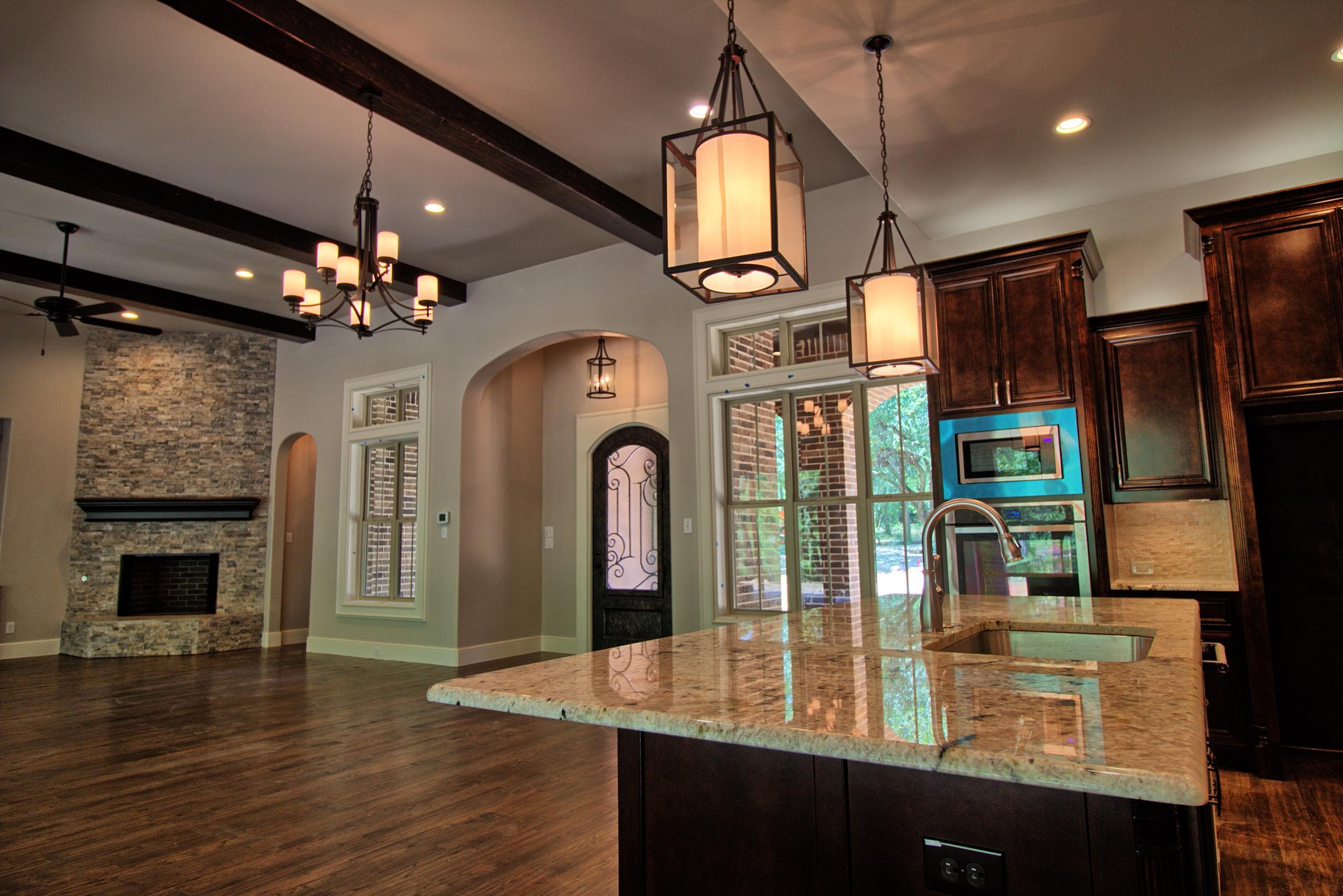 Kitchen and living area with granite island, lantern pendants, dark beams, hardwood floors, stacked stone fireplace and arched iron and glass door