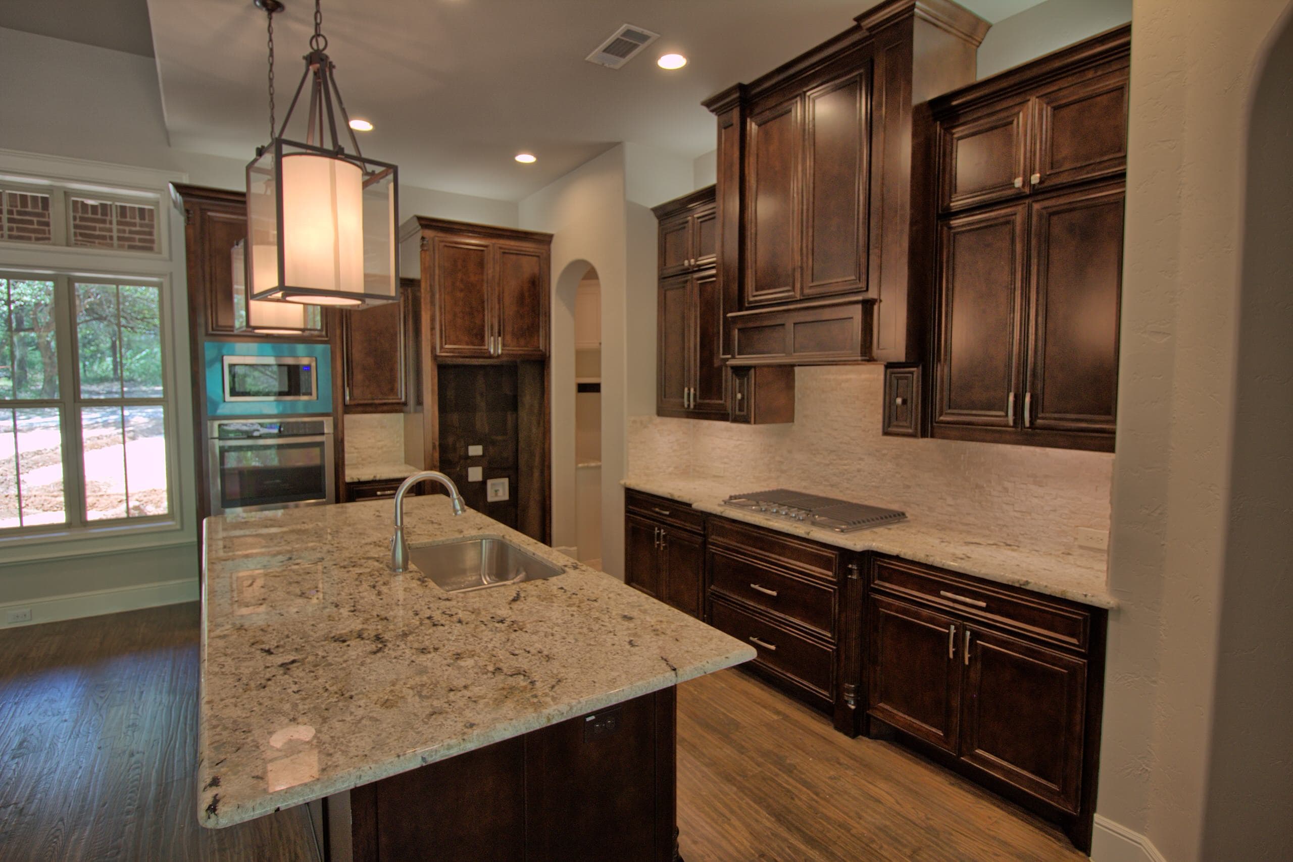 Kitchen with dark stained cabinets, granite island with undermount sink, stainless gas cooktop, white stacked stone backsplash and lantern pendant