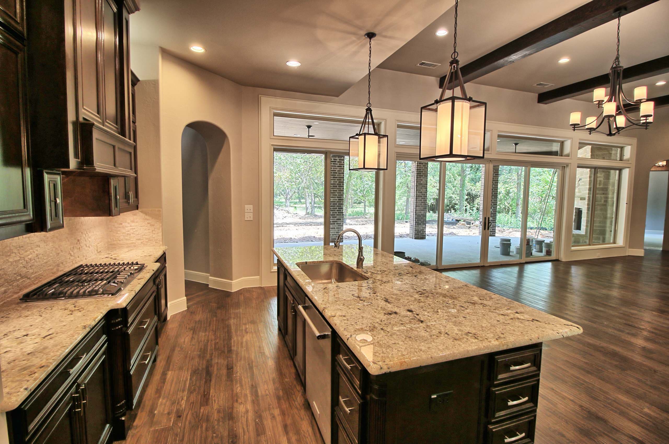 Kitchen with dark cabinets, granite counters, island sink, lantern pendants, wood ceiling beams, hardwood floors and glass doors to a covered patio