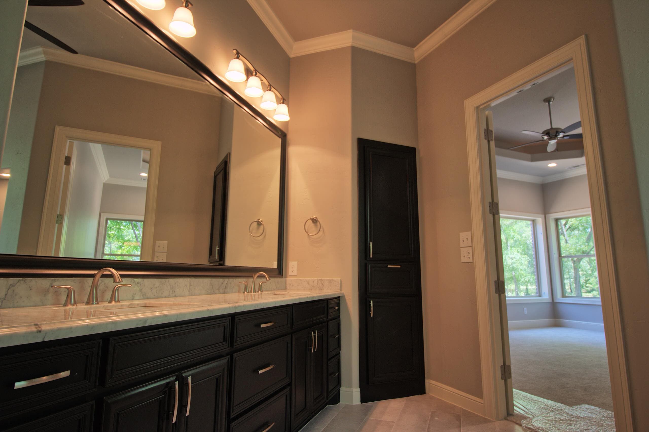 Bathroom with dark double vanity, white marble top, bell-shade light bar, tall linen cabinet, tile floor and doorway to a carpeted bedroom