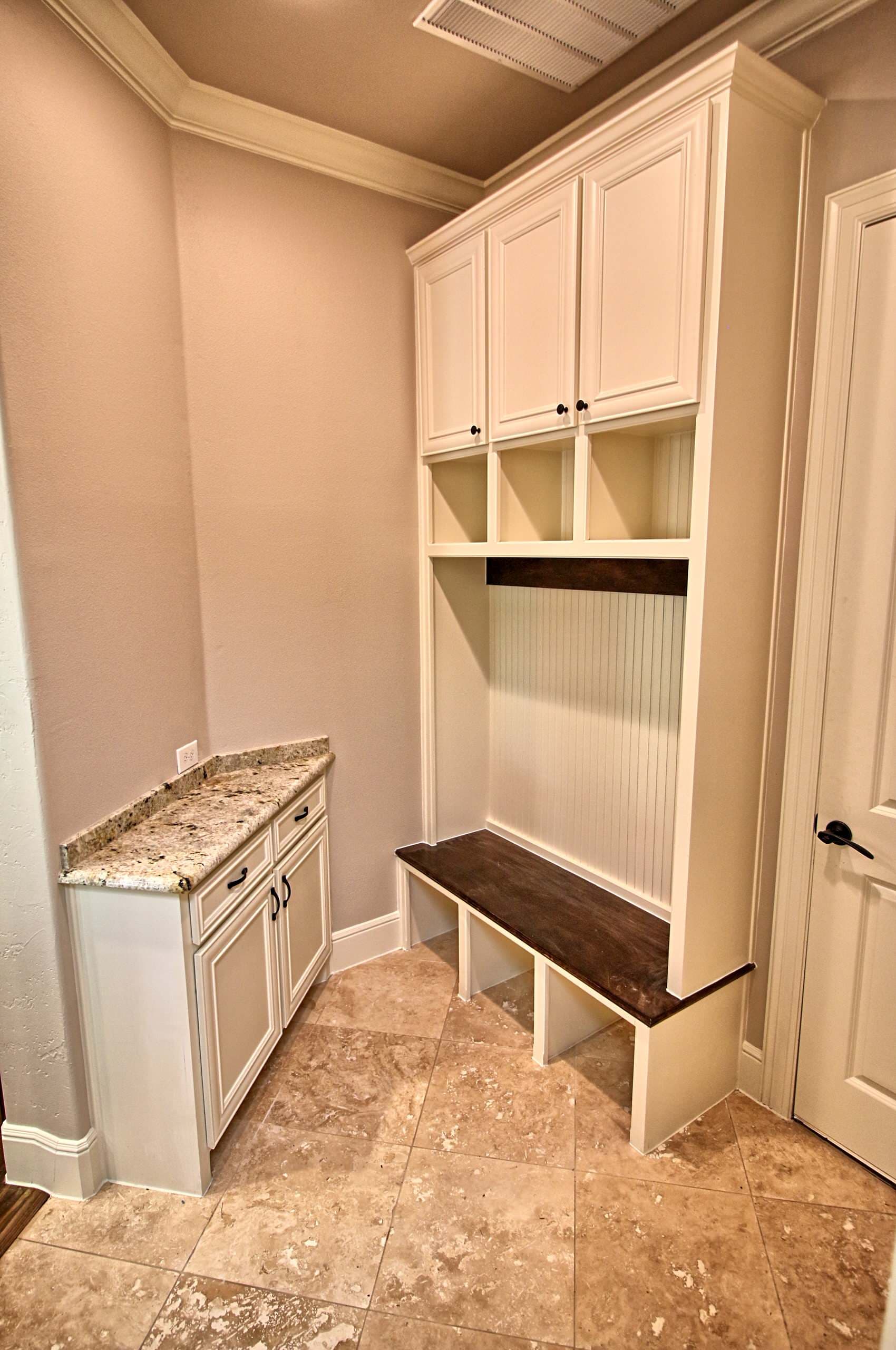 Mudroom with cream built-in locker bench, beadboard back, upper cabinets, stained wood seat, granite-topped cabinet and diagonal travertine tile floor