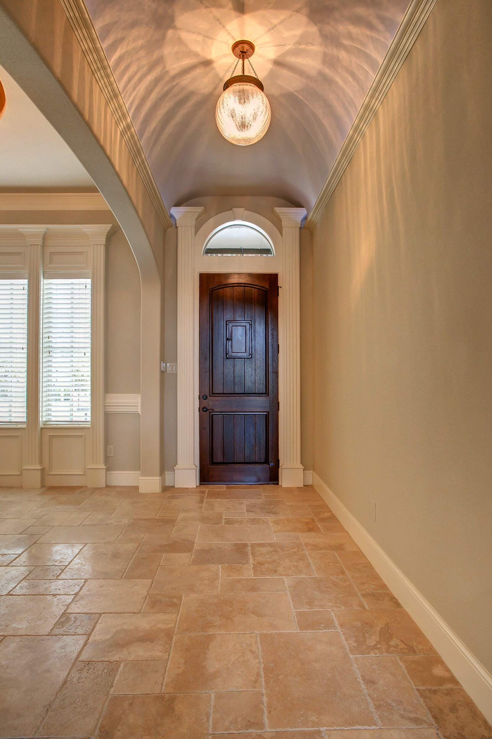 Entry with barrel-vaulted ceiling, glass globe pendant, travertine floor and stained wood front door with arched transom between fluted pilasters