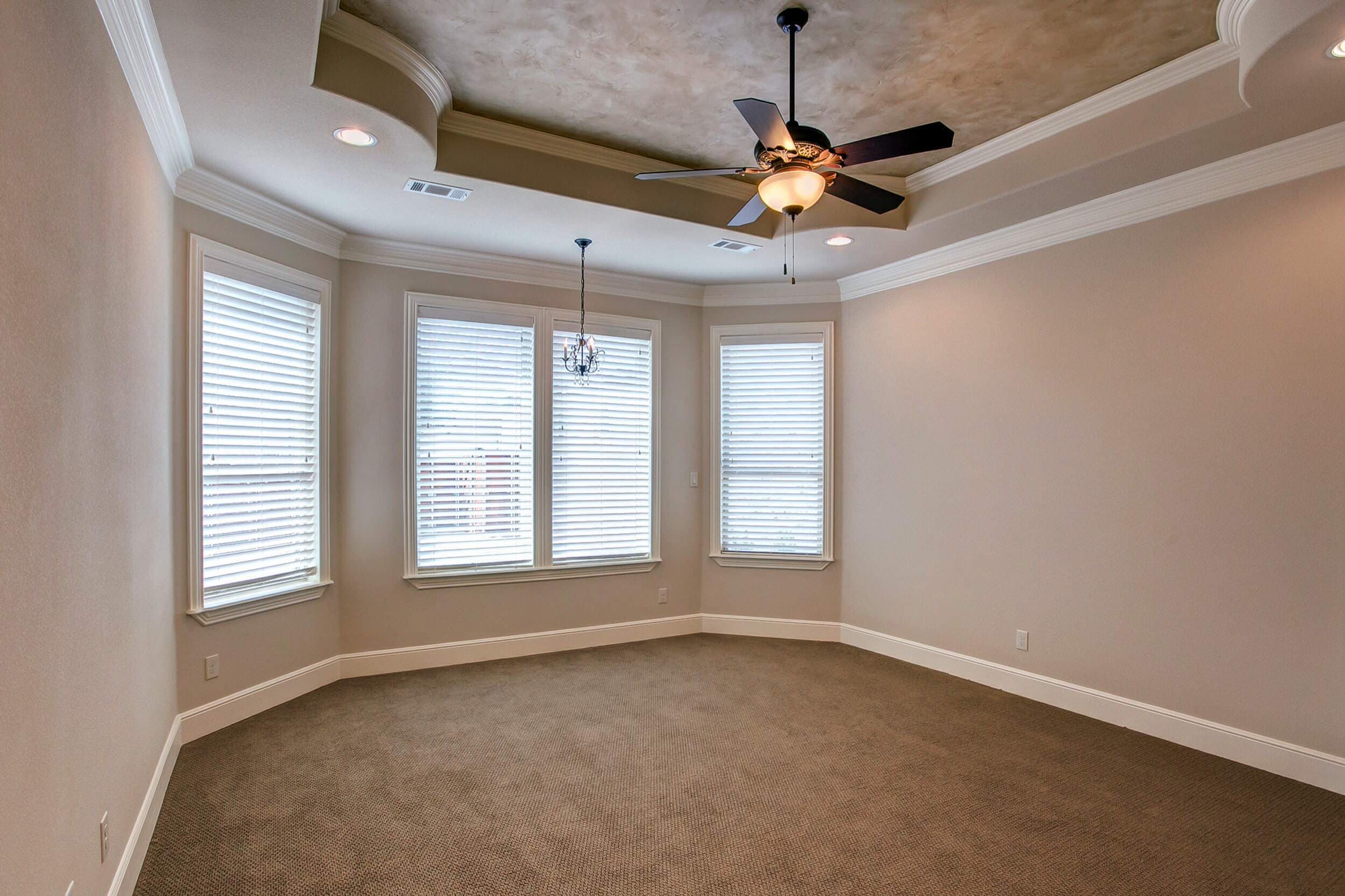 Bedroom with bay window sitting area, small chandelier, stepped tray ceiling with faux finish, ceiling fan, crown molding and brown carpet
