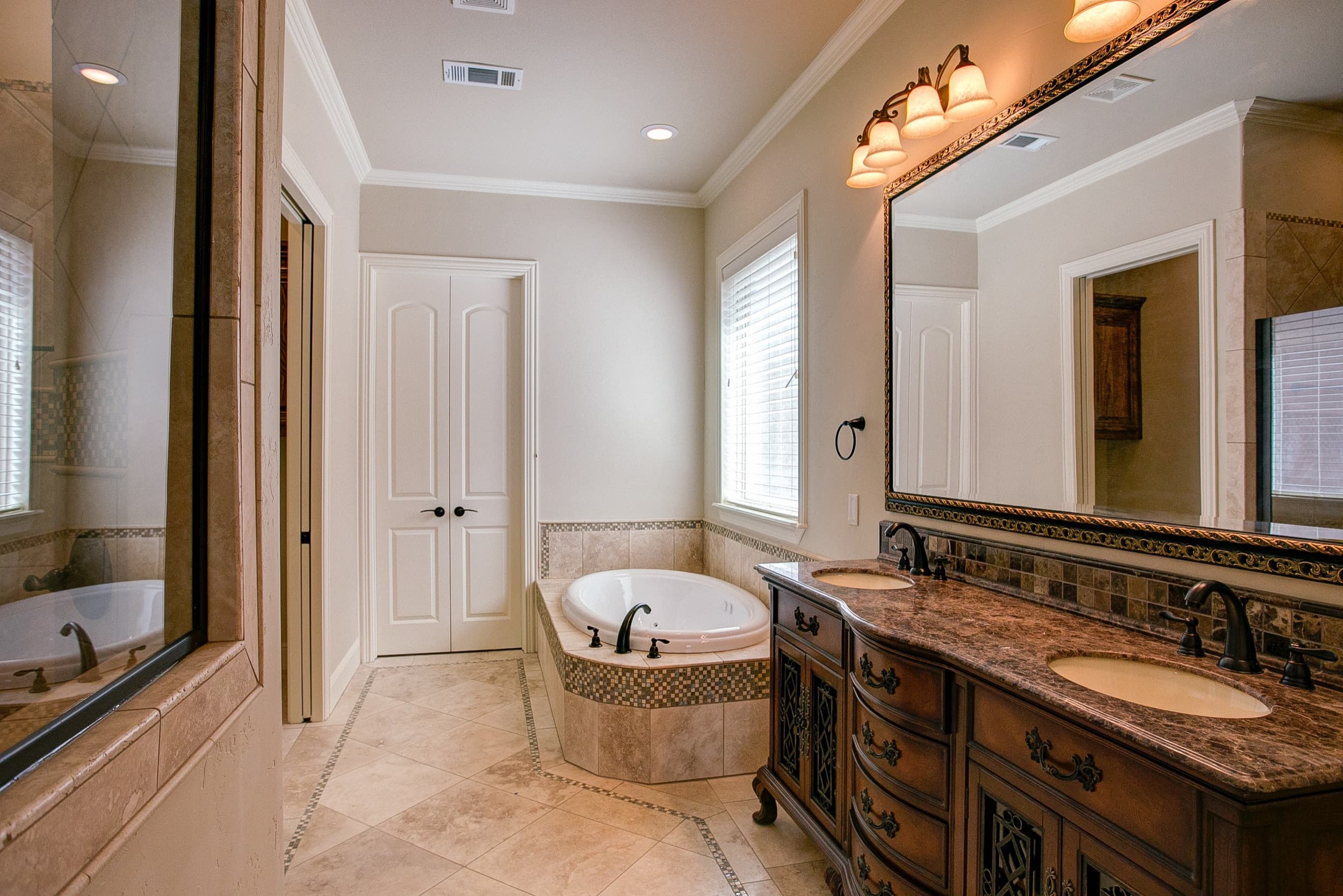 Bathroom with corner jetted tub in tile and mosaic surround, furniture-style double vanity, ornate framed mirror, bronze fixtures and diagonal tile floor