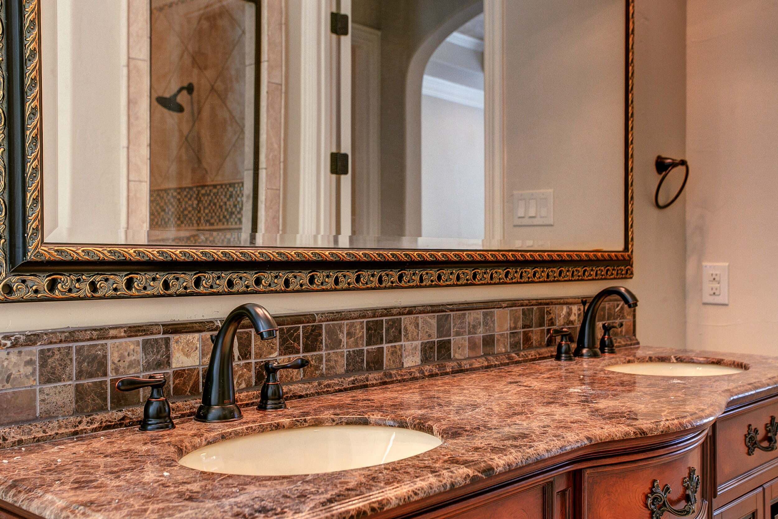 Double vanity with dark marble countertop, oval undermount sinks, oil-rubbed bronze faucets, marble mosaic backsplash and ornate black and gold mirror frame