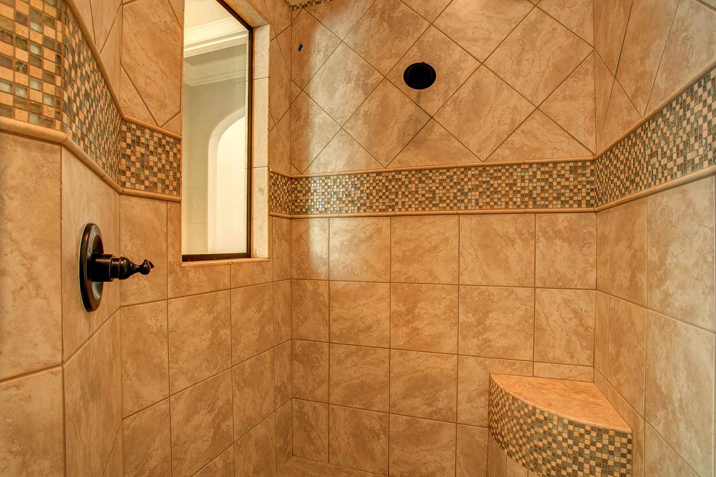 Walk-in shower with diagonal beige tile above an iridescent glass mosaic band, corner bench, bronze valve trim and interior window