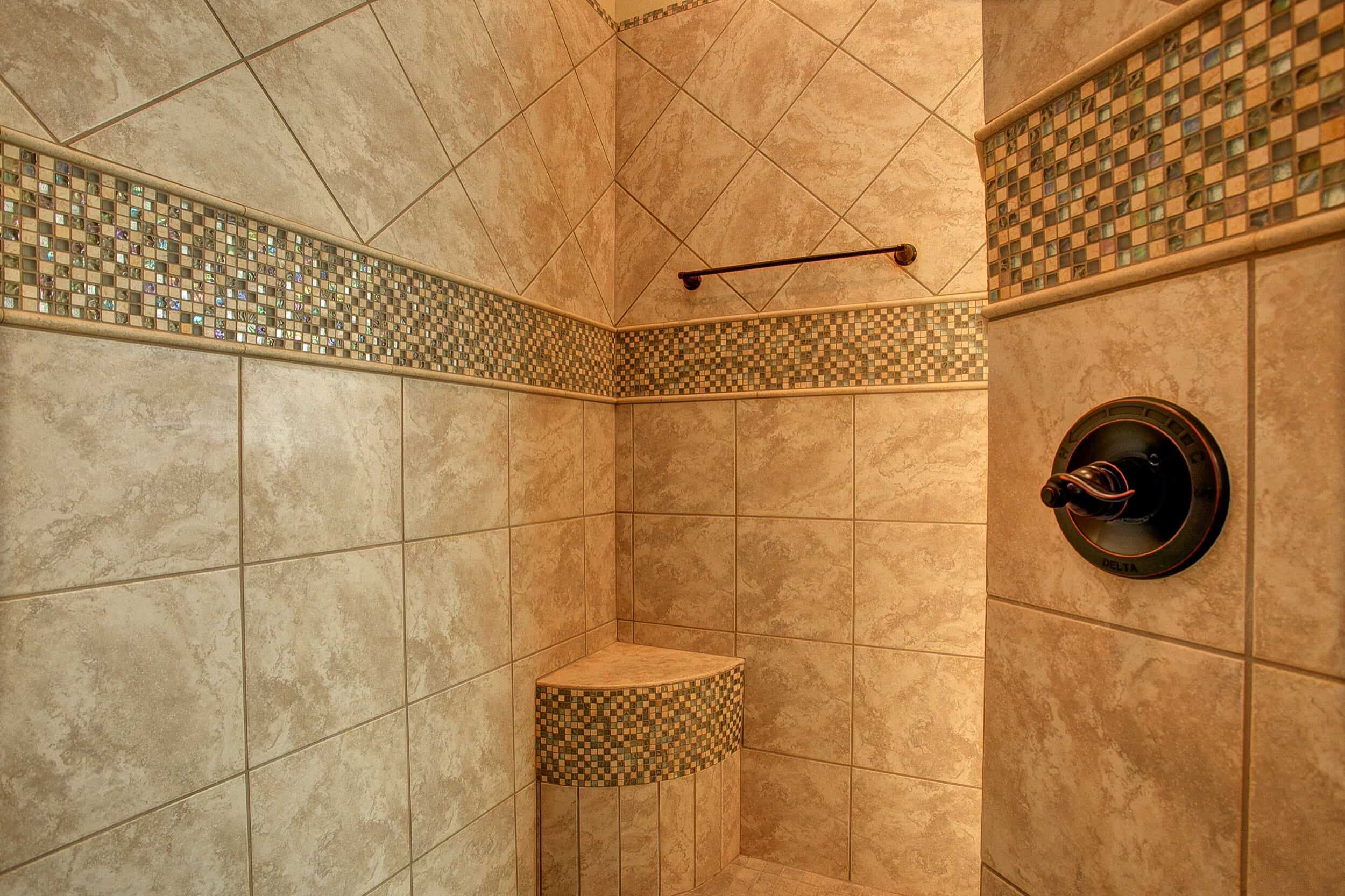 Shower corner with beige marble-look tile, iridescent glass mosaic band, mosaic-faced corner bench, bronze towel bar and bronze valve trim