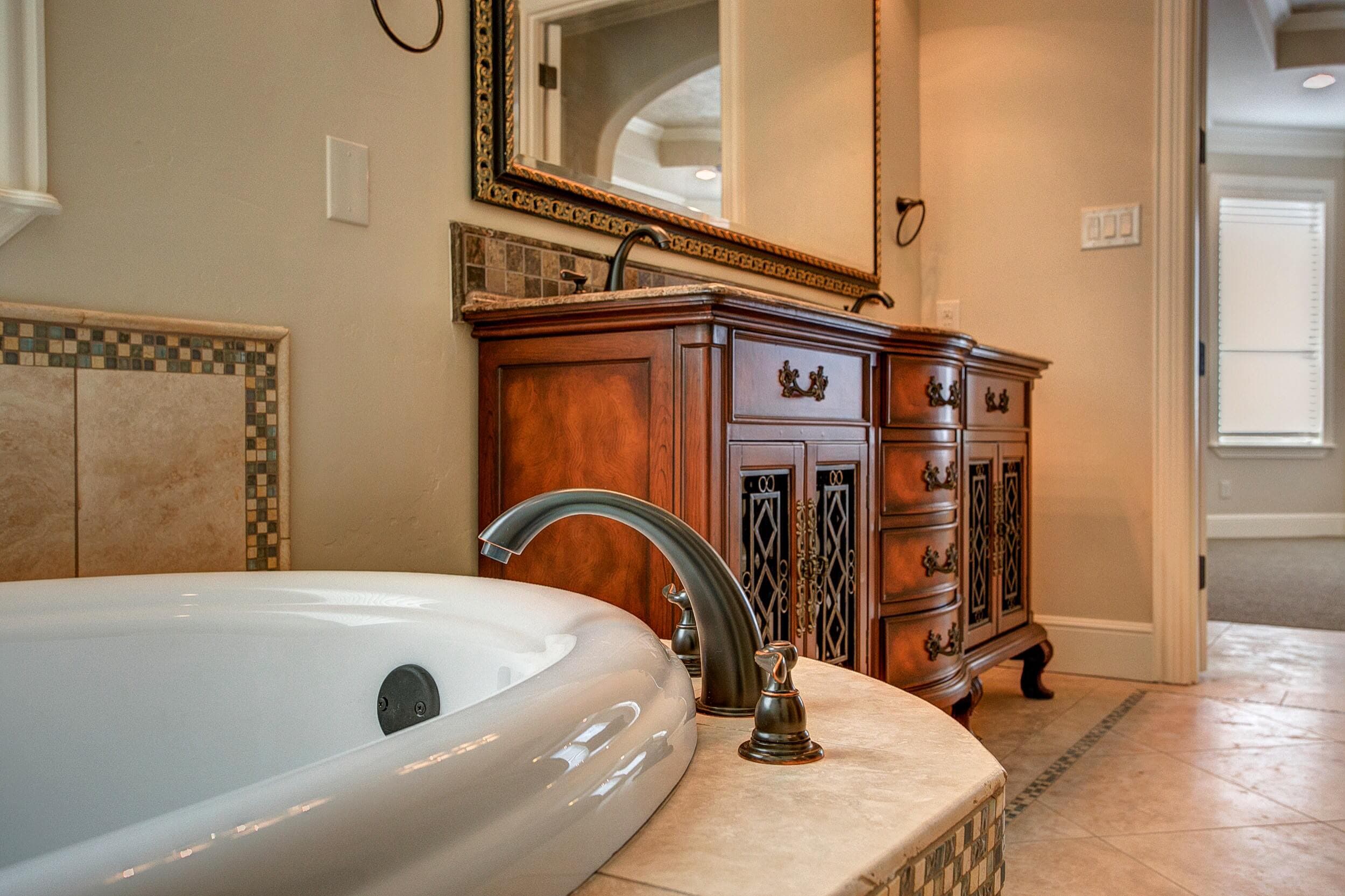 Jetted tub with oil-rubbed bronze faucet and stone deck, facing a furniture-style wood double vanity with iron grille doors