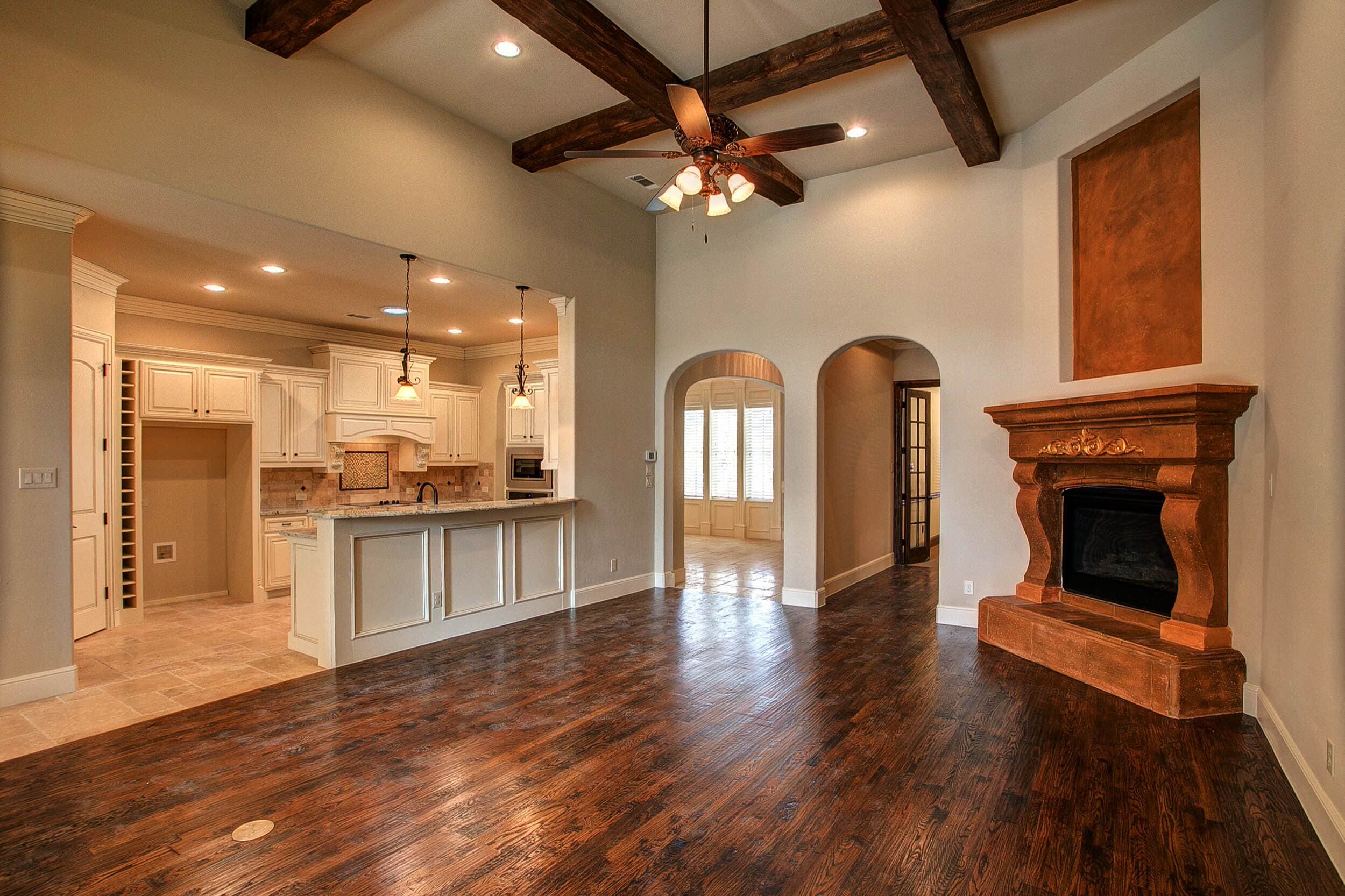 Living room with hand-scraped hardwood floors, dark ceiling beams, ornate corner fireplace, arched doorways and a wide opening to the kitchen
