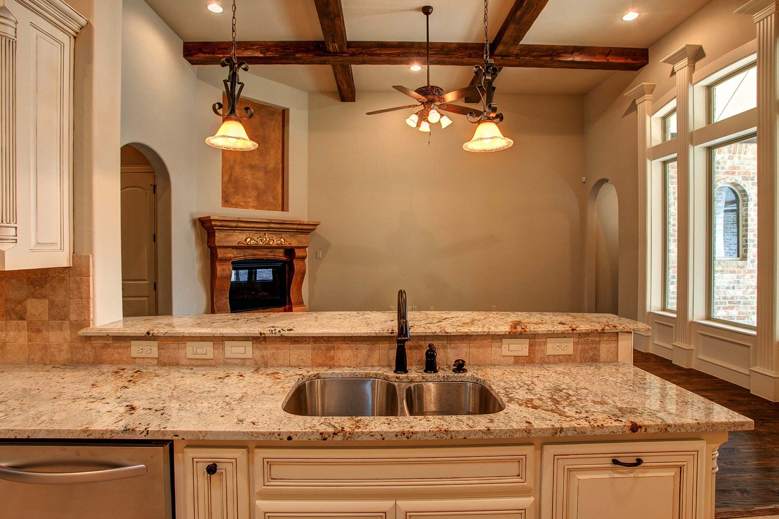 Kitchen island with granite countertop, double stainless sink, bronze faucet and wrought iron pendants, facing a beamed living room with corner fireplace