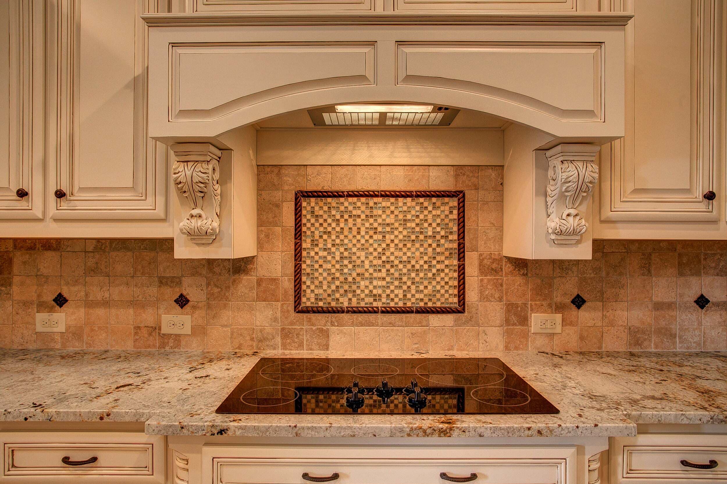 Black glass cooktop under an arched hood with carved corbels, cream glazed cabinets, tumbled travertine backsplash with framed mosaic and granite counters