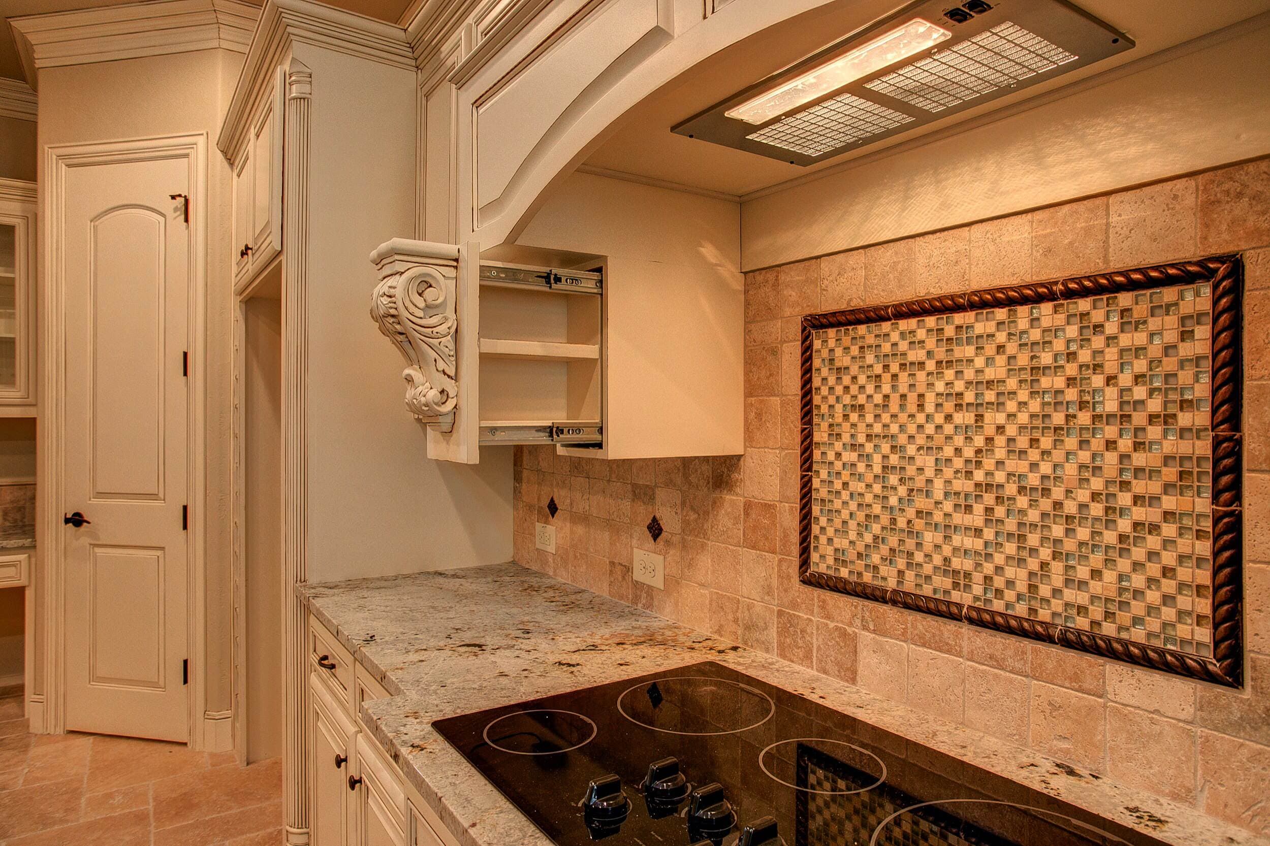 Cooktop area with cream glazed cabinets, open pull-out spice rack with carved corbel, granite counters and travertine backsplash with framed mosaic