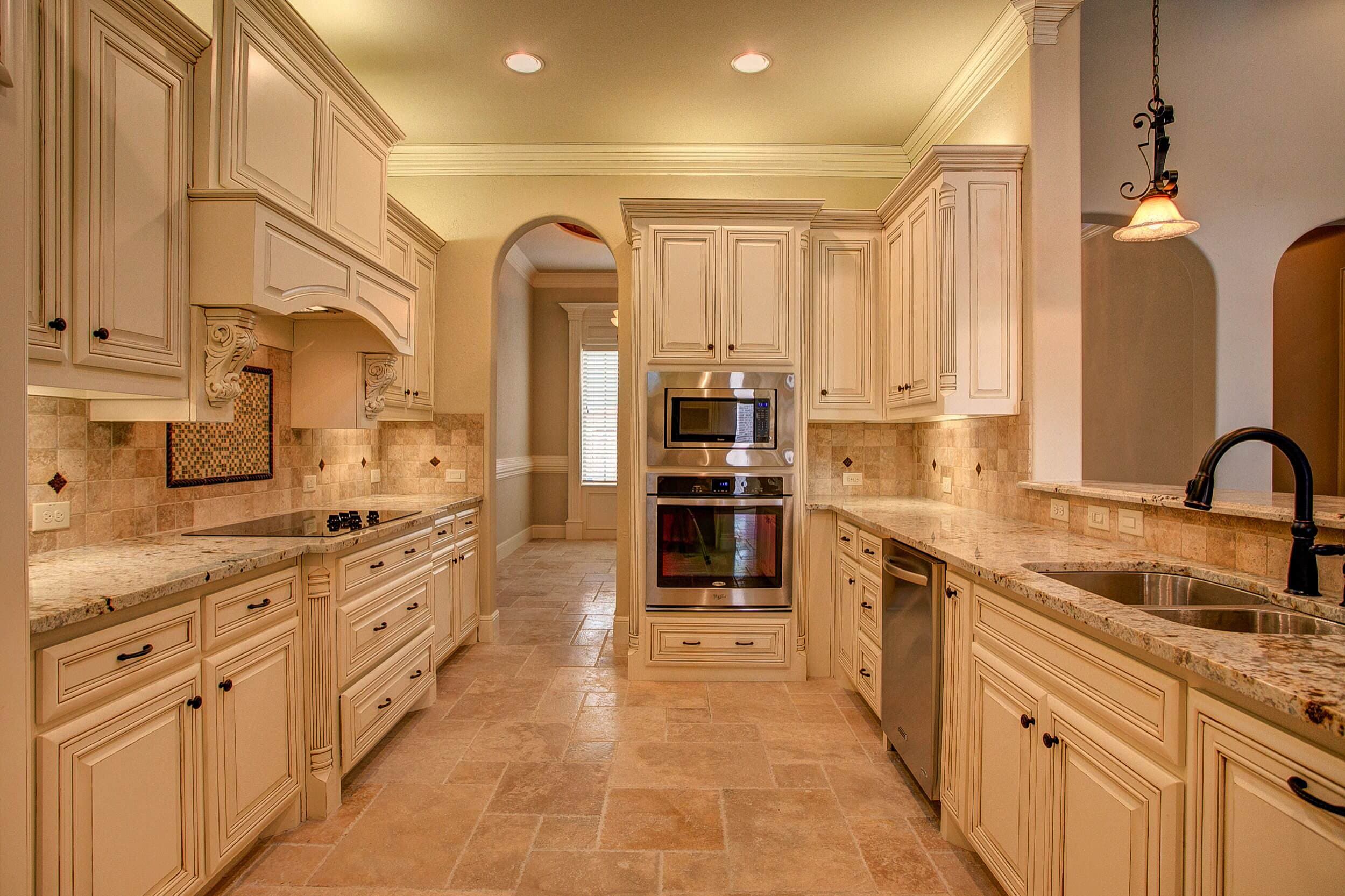 Kitchen with cream glazed cabinets, granite counters, travertine floor and backsplash, stainless wall oven and microwave, and a central arched doorway