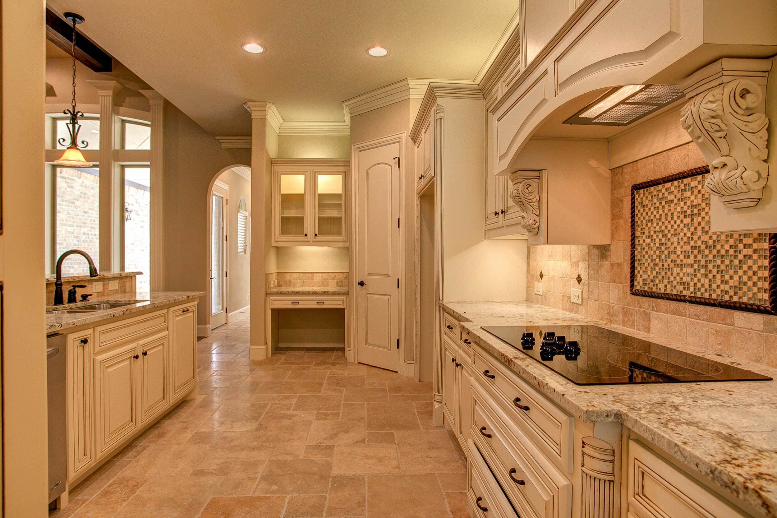 Kitchen with cream glazed cabinets, granite counters, carved corbels and travertine floor leading to a built-in desk with lit glass cabinets