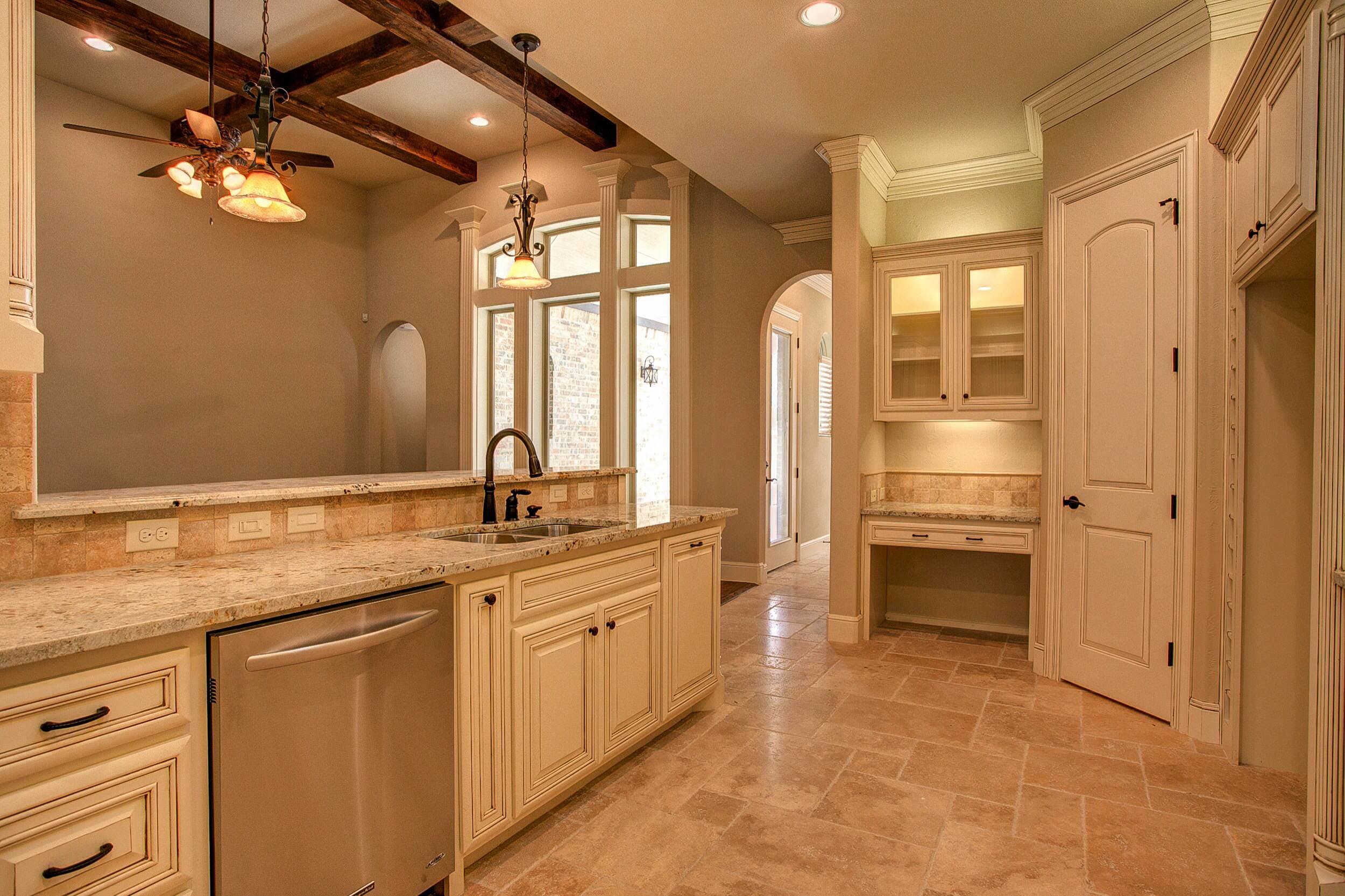 Kitchen sink run with cream glazed cabinets, granite counters, raised bar, stainless dishwasher and travertine floor, open to a beamed living room