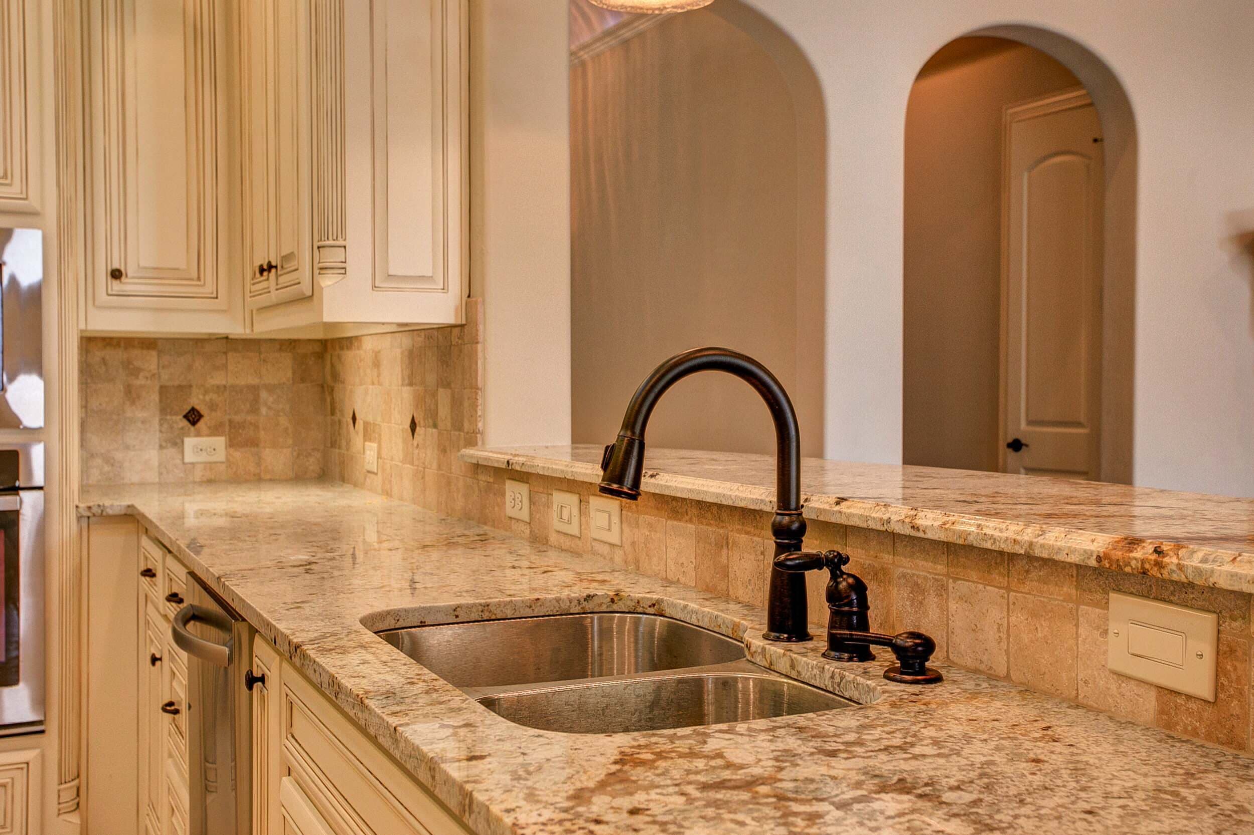 Double stainless undermount sink with oil-rubbed bronze pull-down faucet in granite counters, travertine backsplash with diamond accents and arched openings beyond