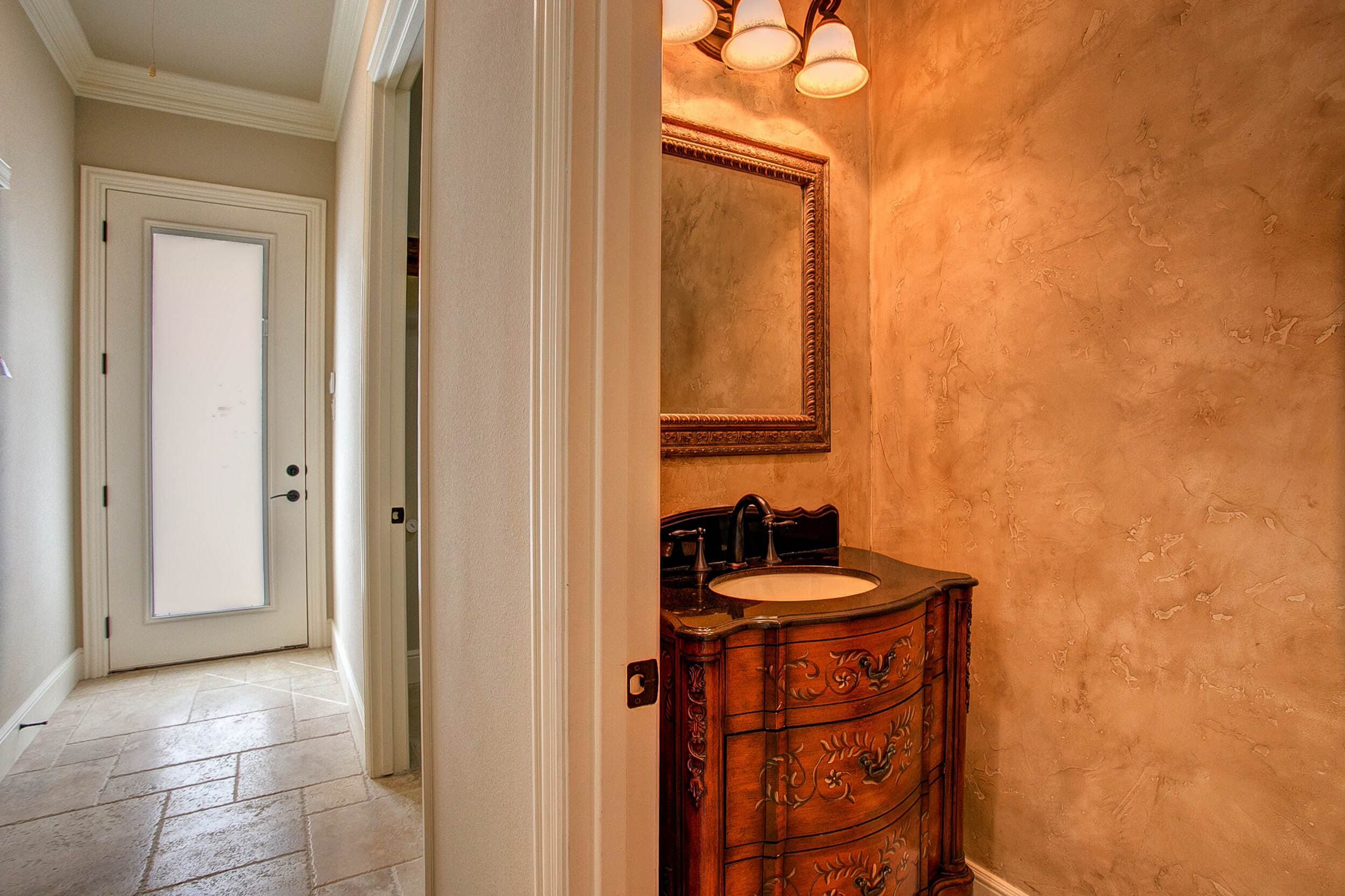 Powder room with furniture-style vanity painted with florals, dark stone top, ornate mirror and faux-finished walls beside a travertine hallway