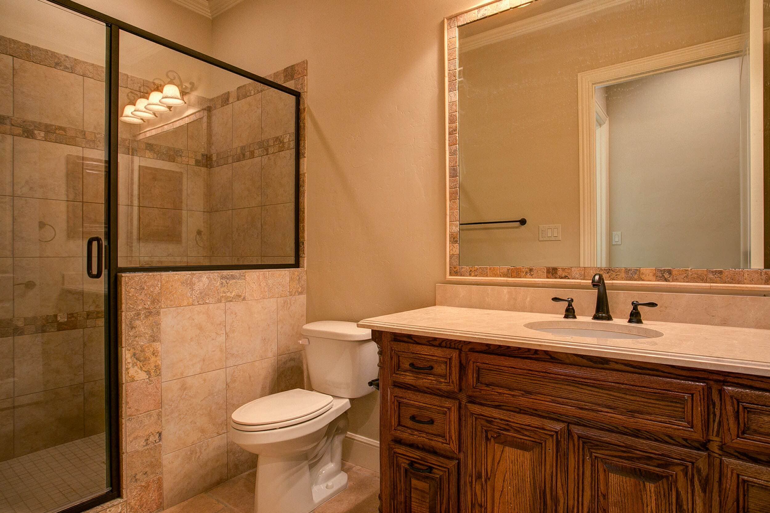Bathroom with stained oak vanity, cream marble top, bronze faucet, mirror framed in tumbled stone tile and a glass-enclosed tiled shower