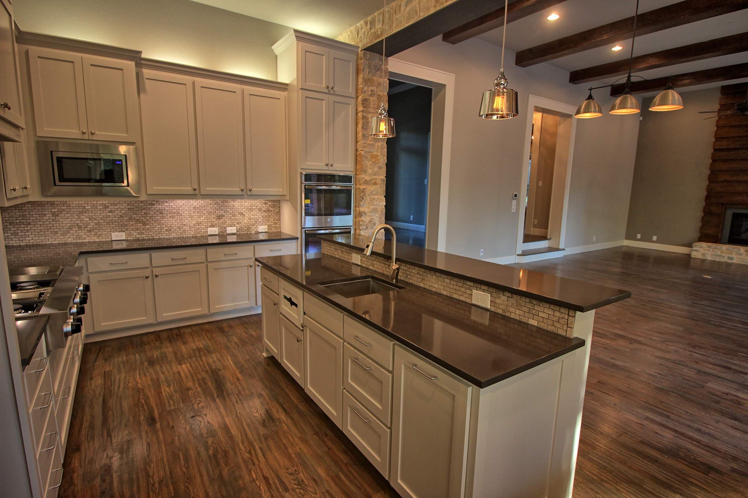 Kitchen with white shaker cabinets, dark countertops, mosaic backsplash, island with raised bar, glass pendants, double wall ovens and wood ceiling beams