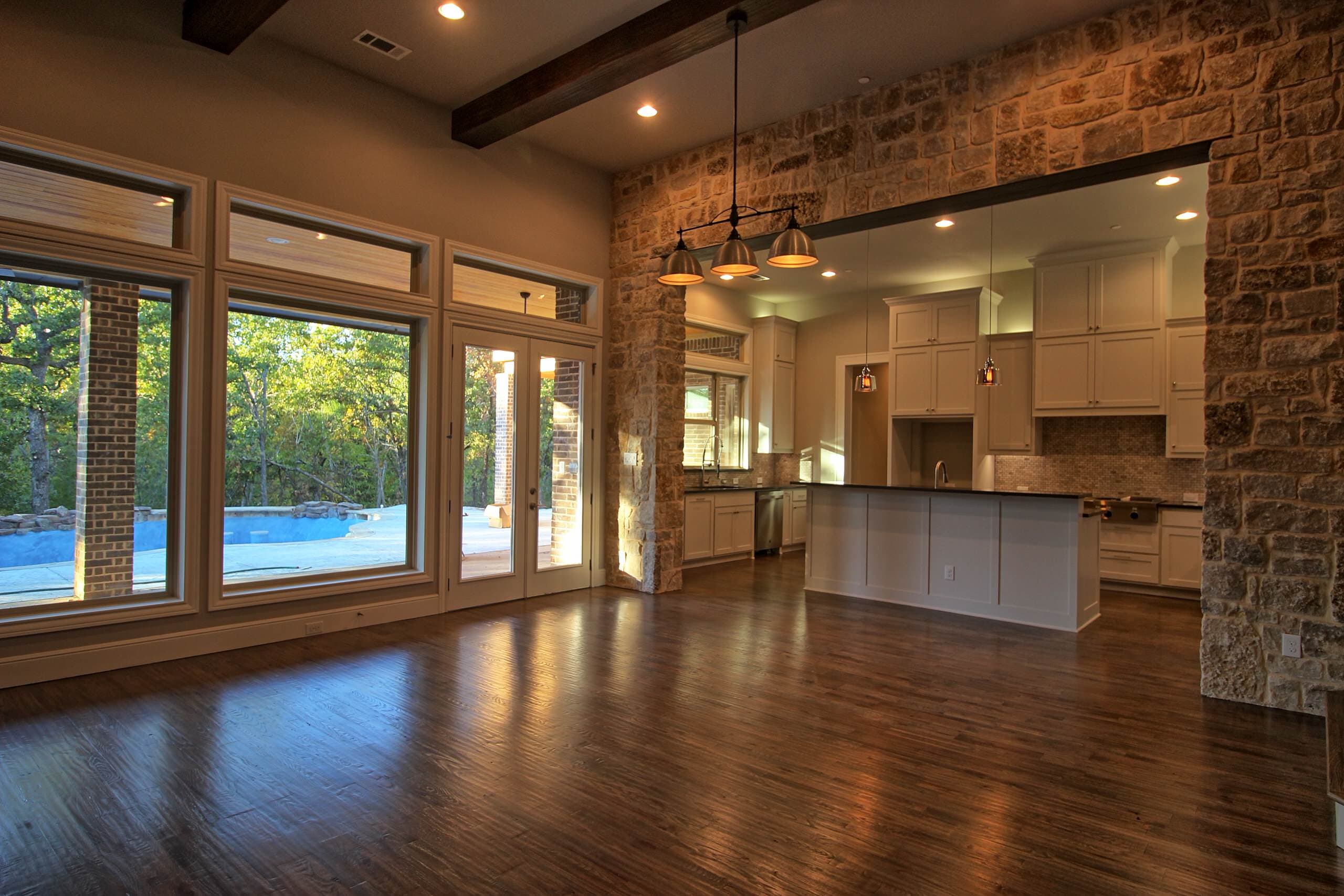 Great room with hardwood floors, a stone wall framing a wide opening to the kitchen, pendant lights and windows overlooking the pool