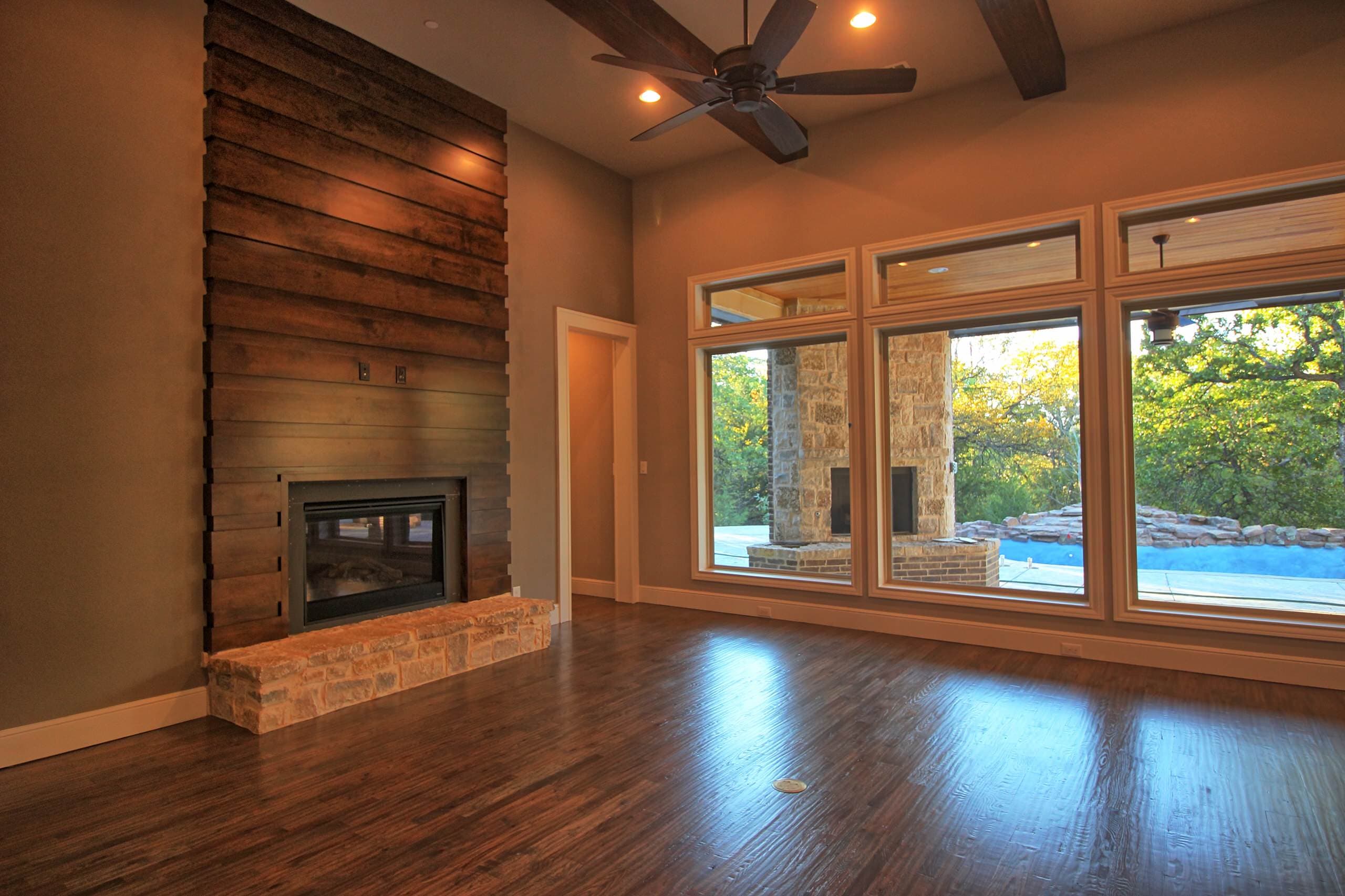 Great room with floor-to-ceiling wood plank fireplace wall, stone hearth, ceiling fan, wood beams and windows facing an outdoor fireplace and pool