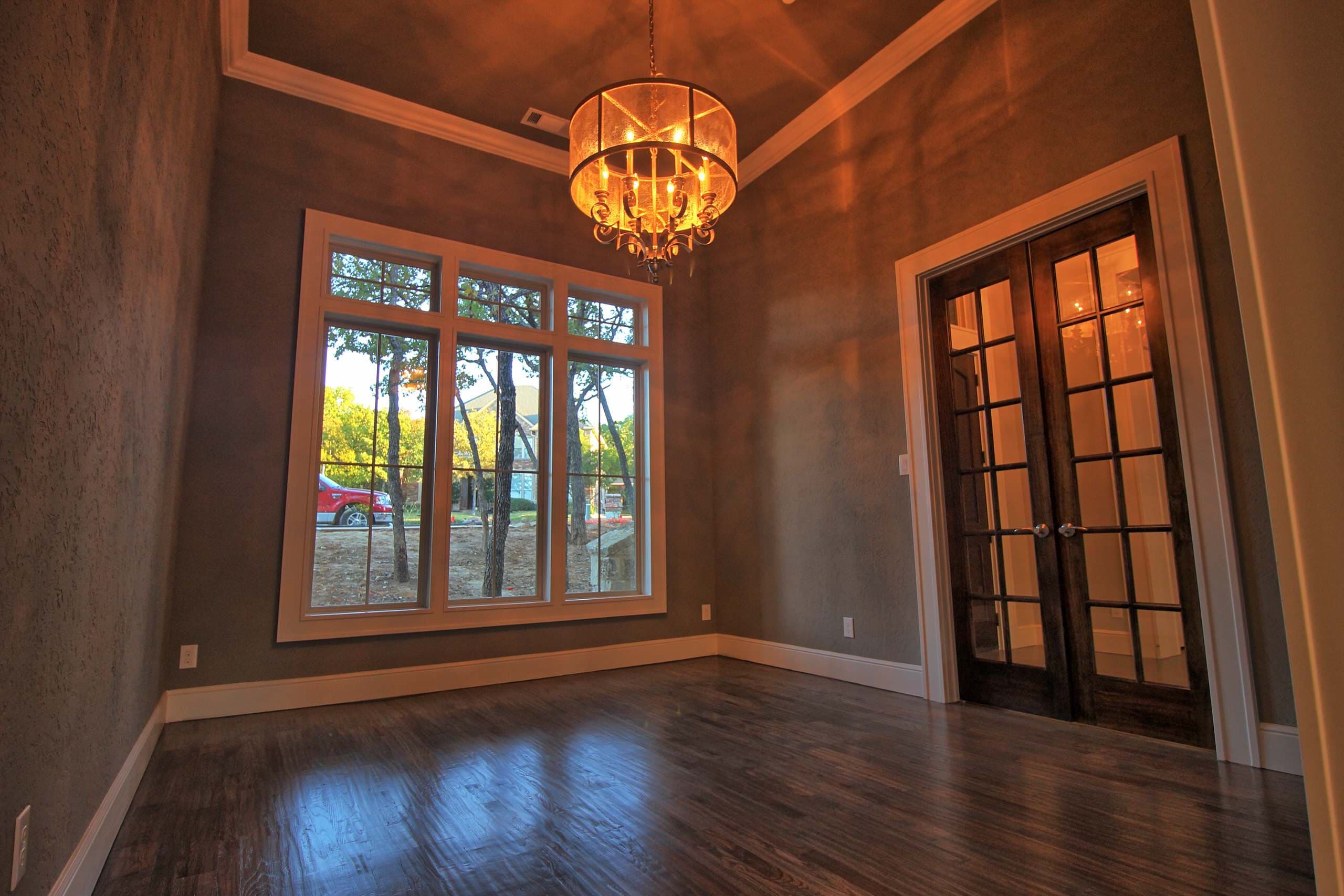 Study with drum chandelier, tray ceiling with crown molding, textured plaster walls, hardwood floors, stained wood French doors and large gridded window