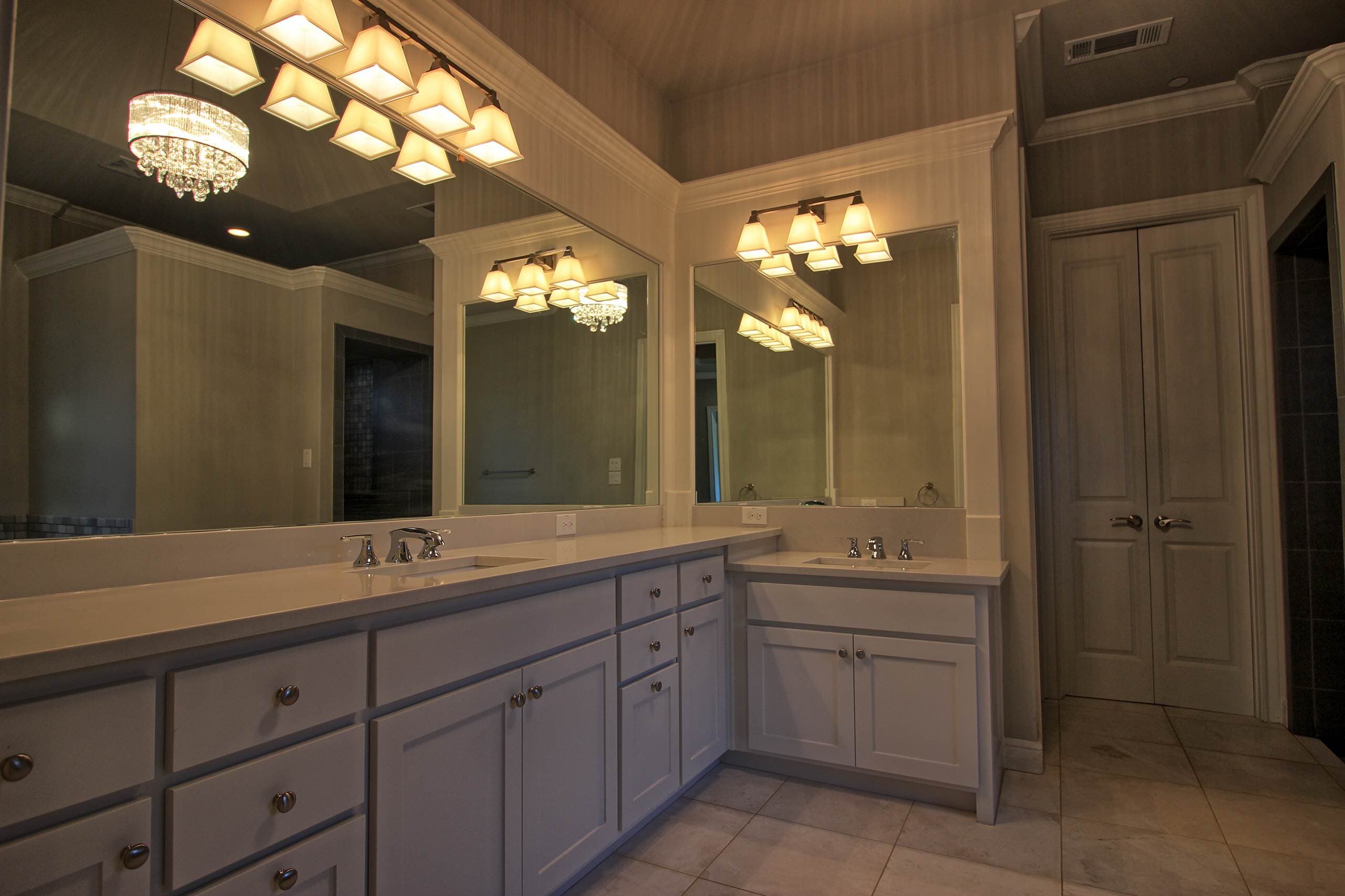 Bathroom with white shaker vanities, white countertops, two sinks, framed mirrors reflecting a crystal chandelier, vanity lights and tile floor