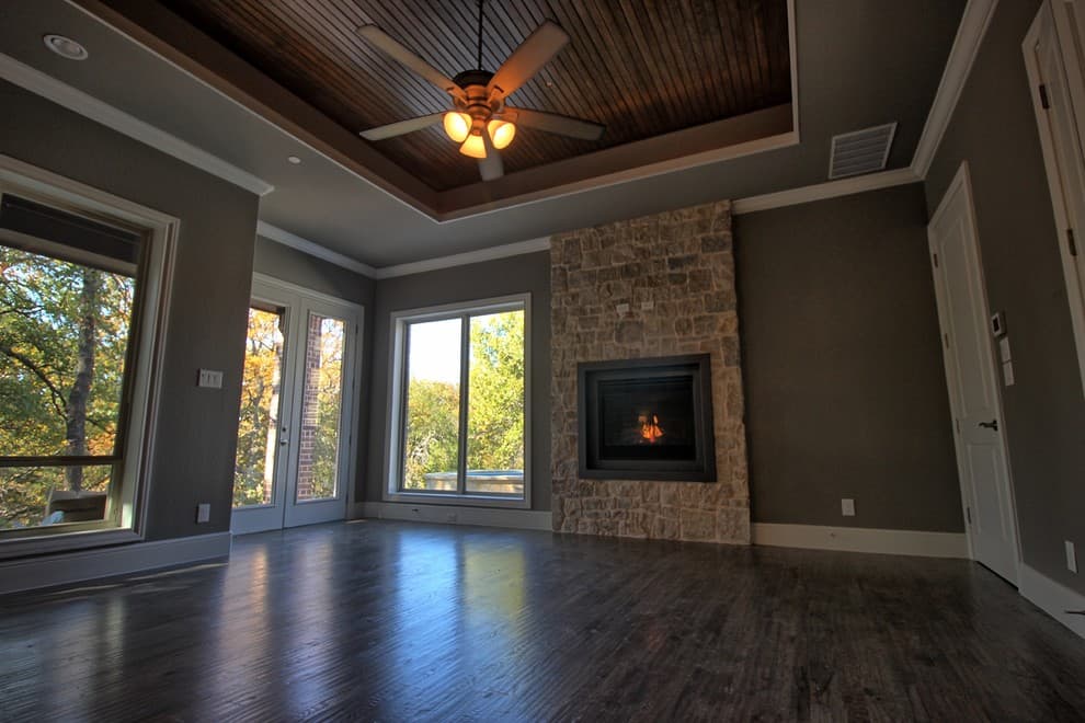 Bedroom with stone fireplace, stained wood plank tray ceiling, ceiling fan, hardwood floors, French doors and windows facing trees