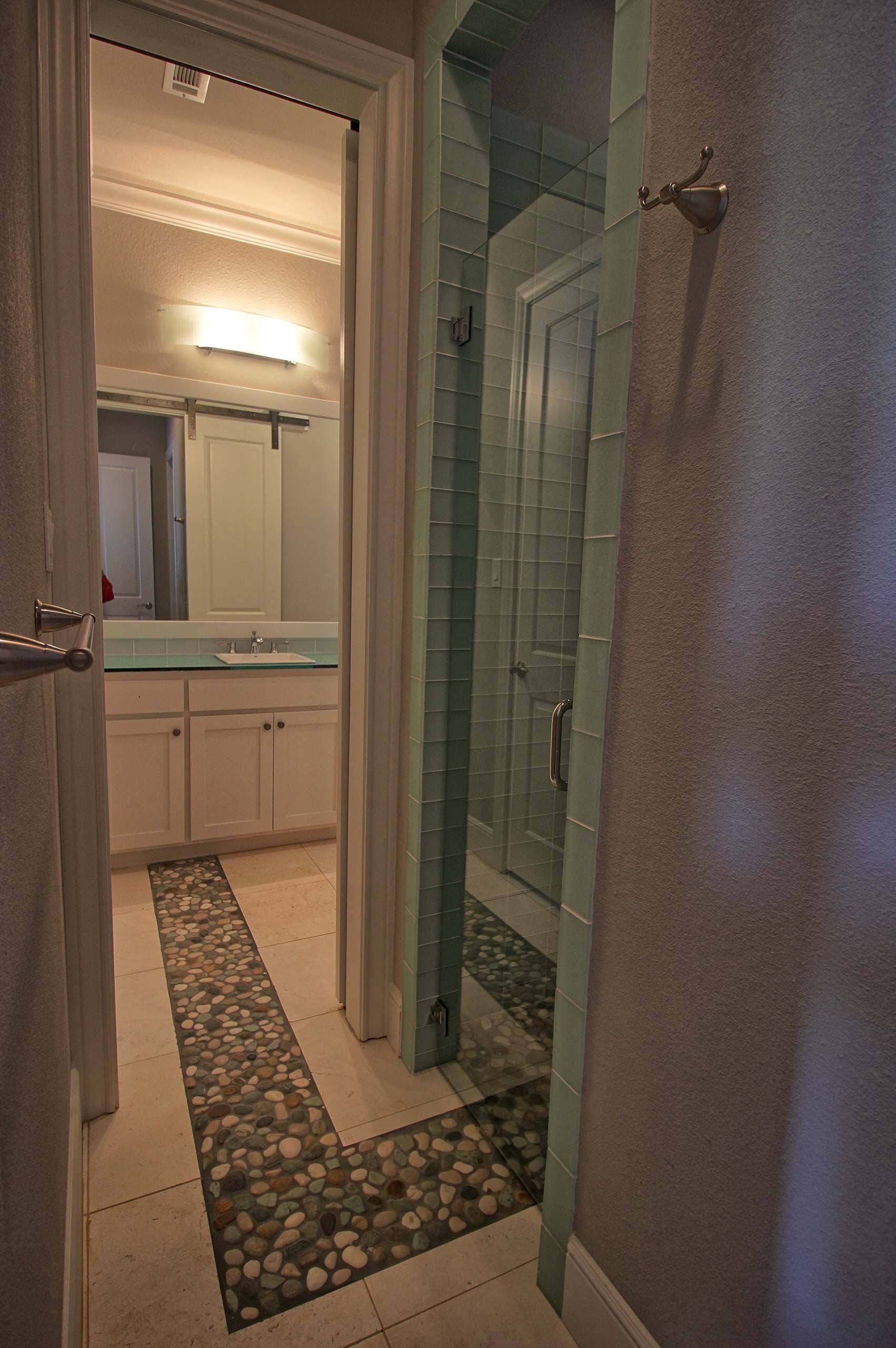 Bathroom with pebble tile runner set into cream floor tile, aqua glass tile shower with frameless door and vanity beyond