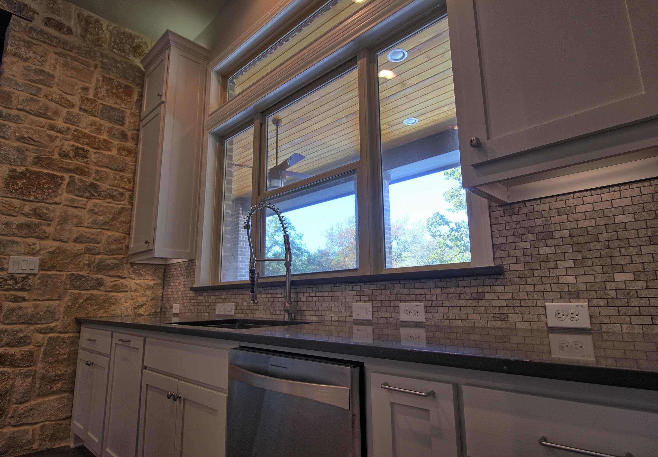 Kitchen sink with commercial-style faucet below a window to the covered patio, dark countertops, white shaker cabinets, mosaic backsplash and stone wall