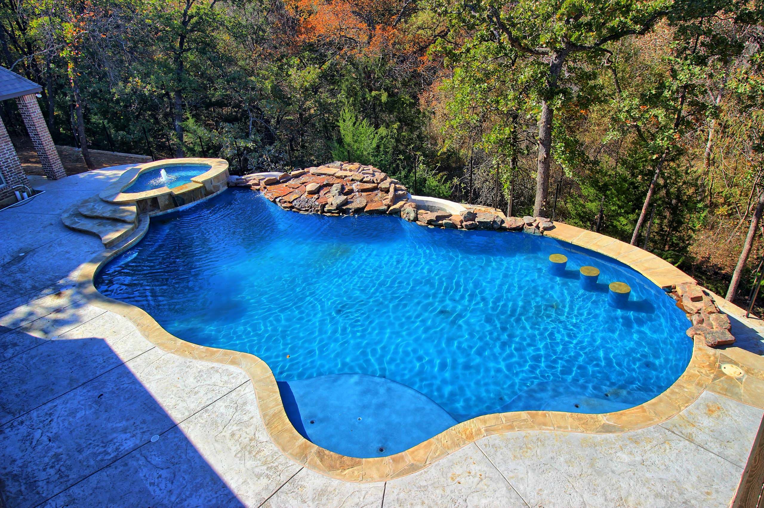 Freeform pool with raised spa, rock waterfall, in-pool stools, sun shelf, flagstone coping and stamped concrete deck beside a wooded hillside
