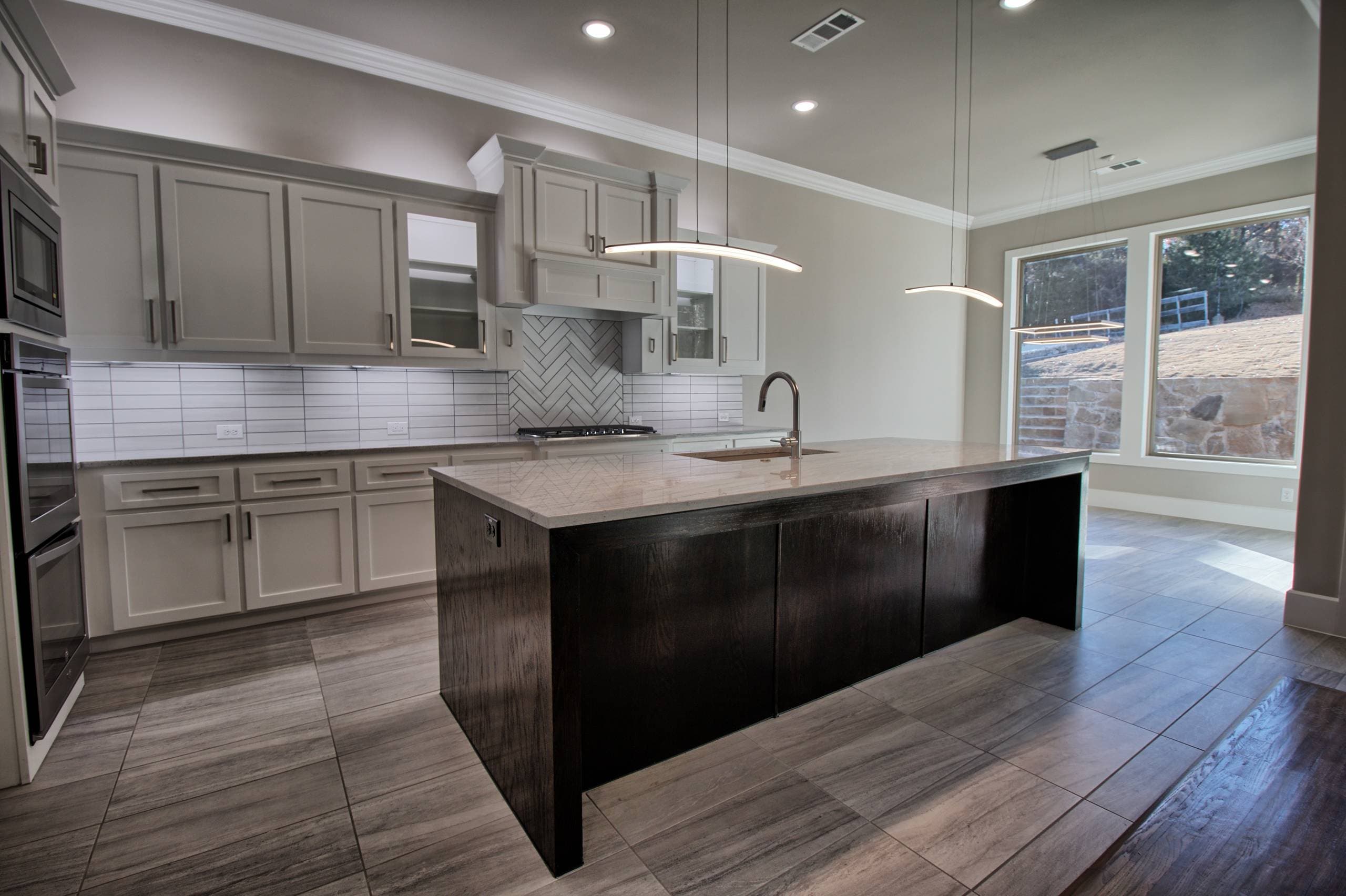 Kitchen with light gray shaker cabinets, herringbone tile accent, dark oak waterfall island, curved LED pendants, and large-format gray tile floor