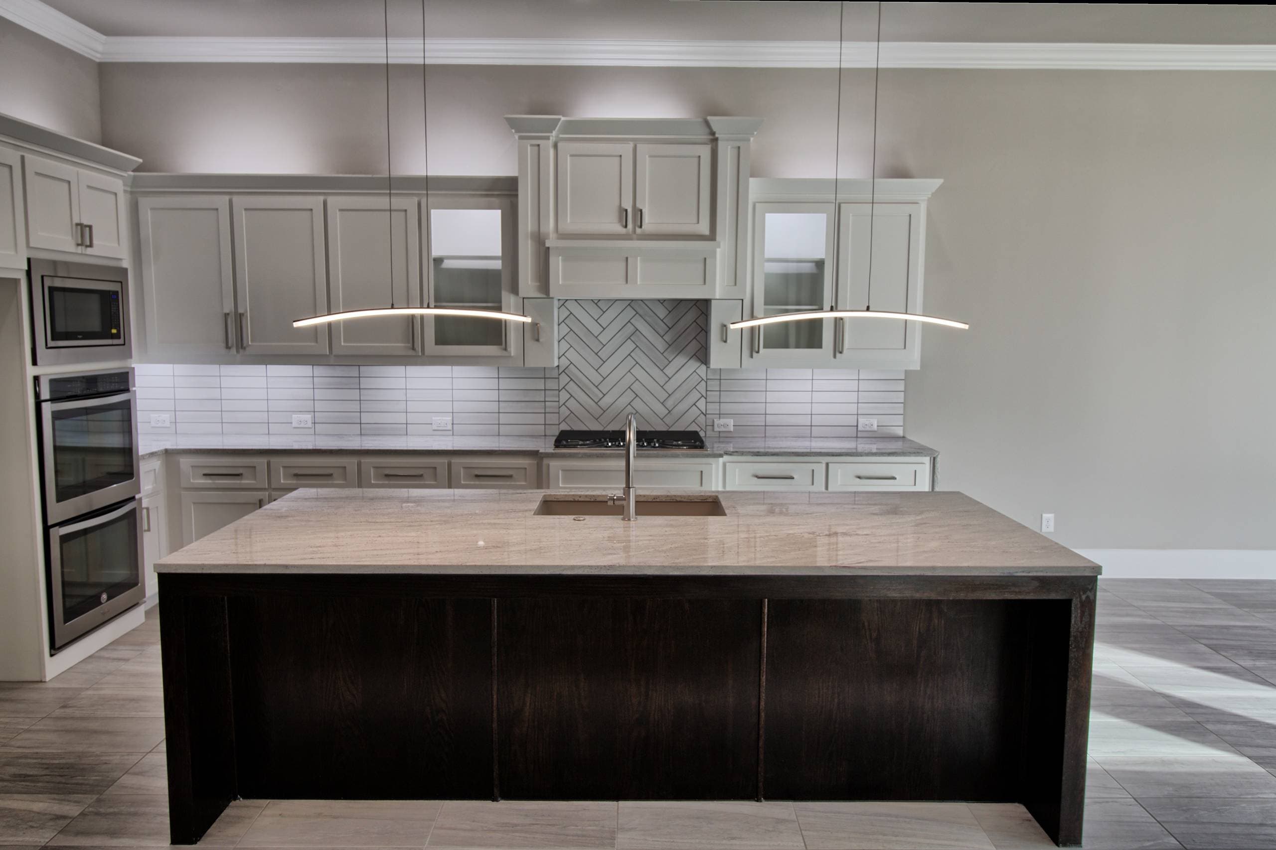 Dark oak kitchen island with granite top and undermount sink under two curved LED pendants, backed by light gray shaker cabinets