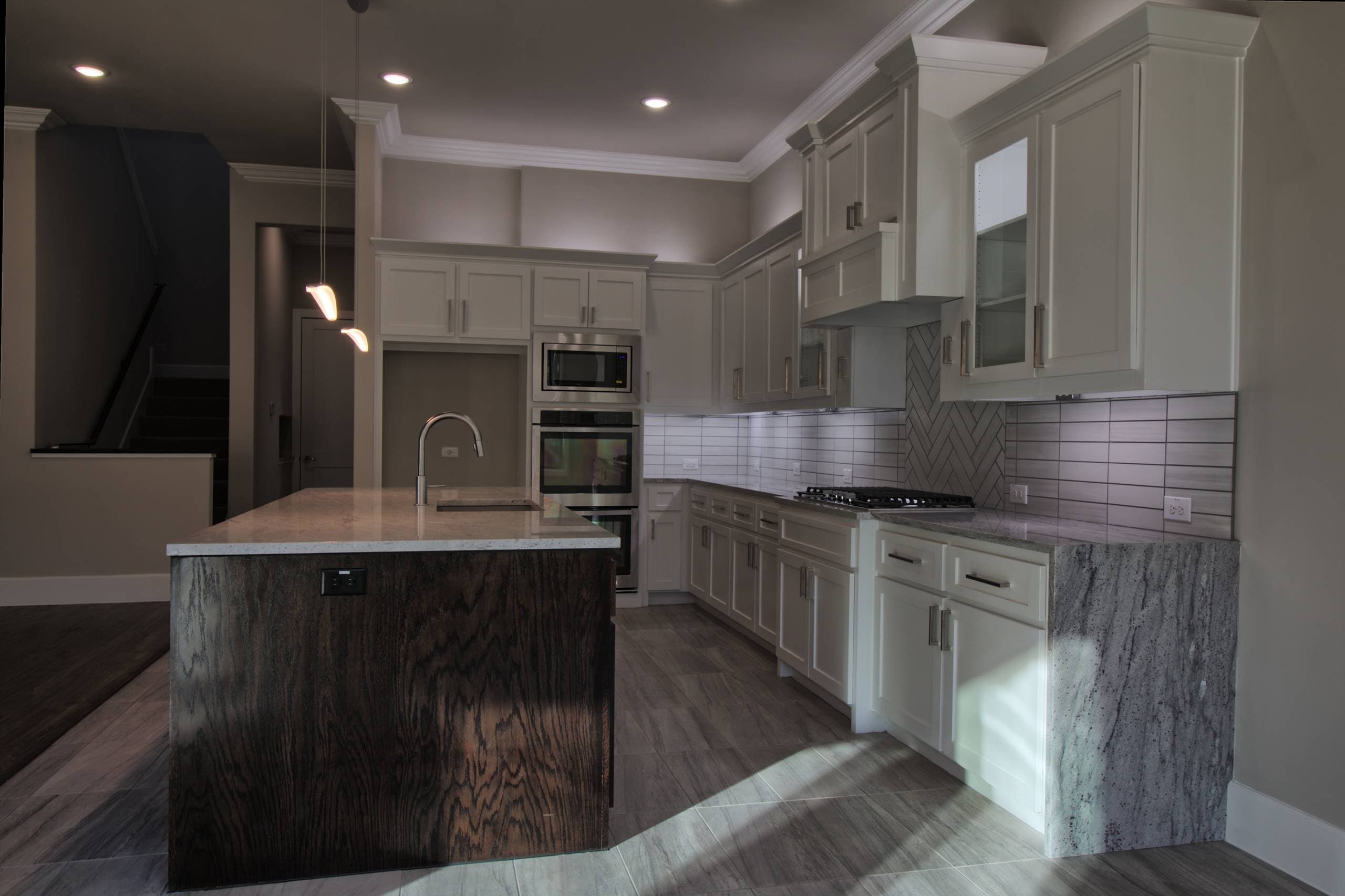 Kitchen with light gray shaker cabinets, granite waterfall counter, dark oak island, herringbone tile behind the gas cooktop, and stainless wall ovens