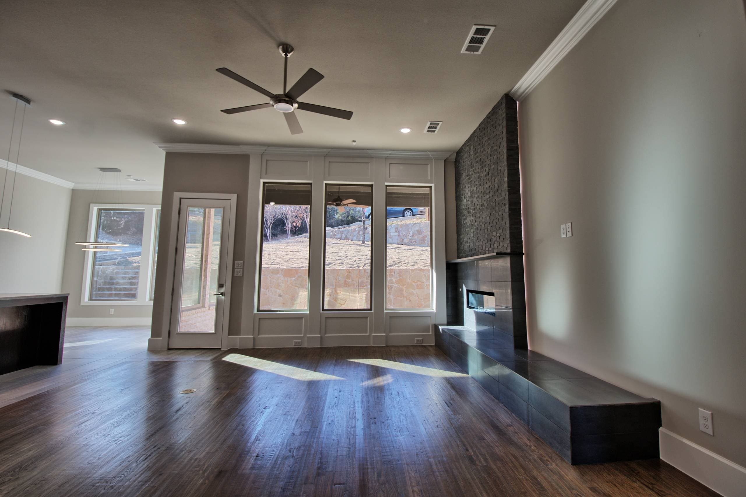 Living room with three tall windows in paneled trim, glass patio door, dark hardwood floor, ceiling fan, and a tiled linear fireplace