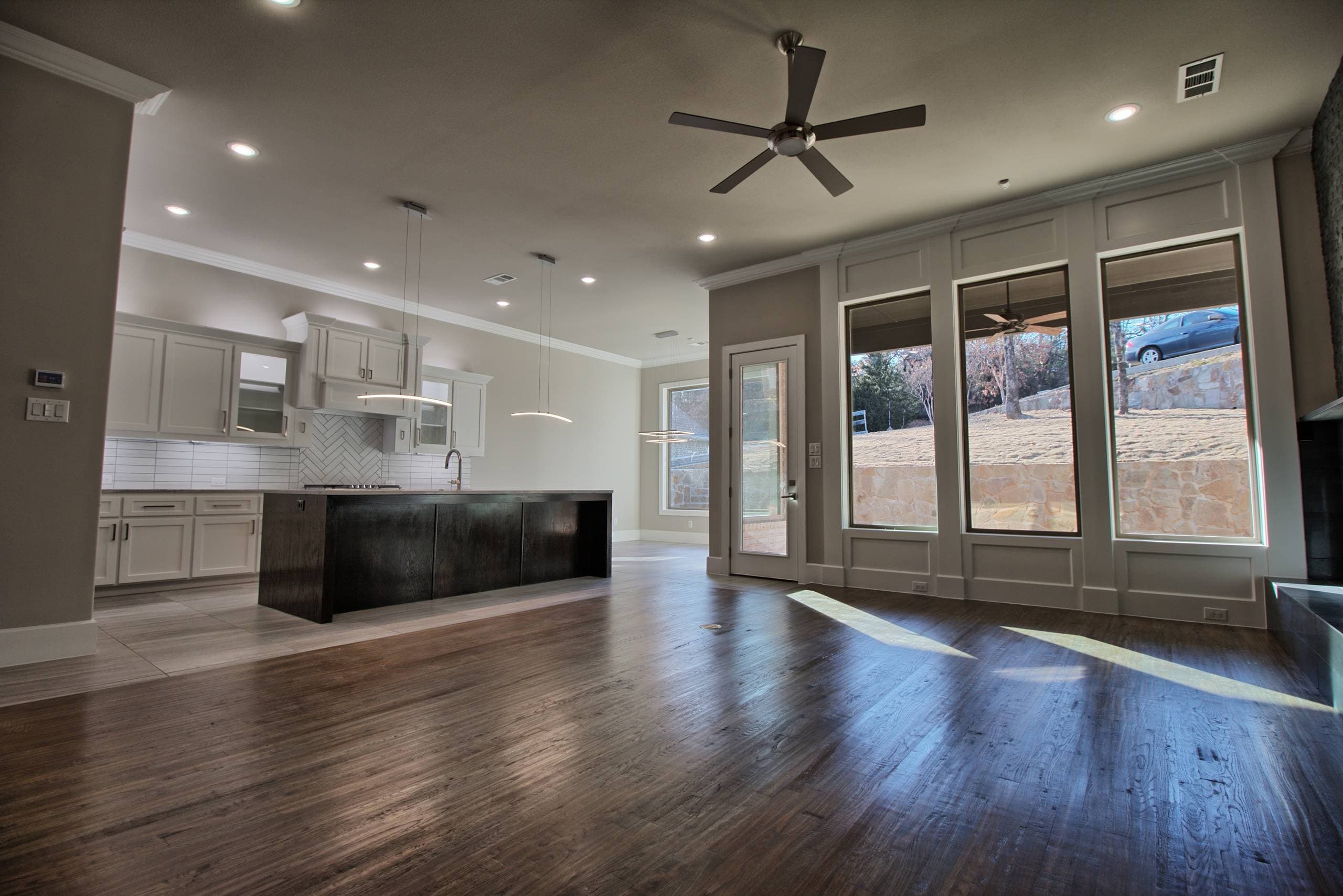 Living room with dark hardwood floor and tall windows, flowing into a kitchen with light gray cabinets and a dark oak island