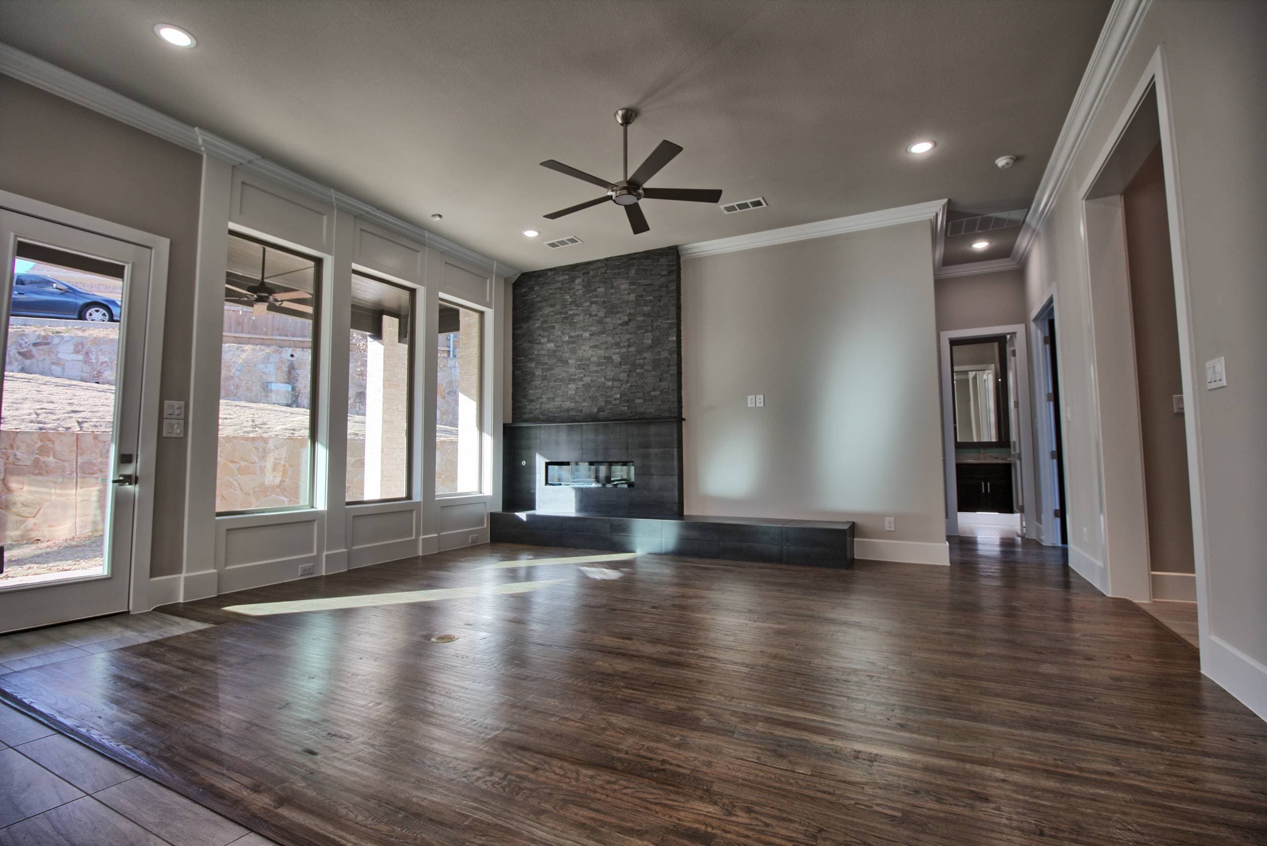 Living room with charcoal stacked stone chimney, linear fireplace over a long tiled hearth, dark hardwood floor, ceiling fan, and tall windows