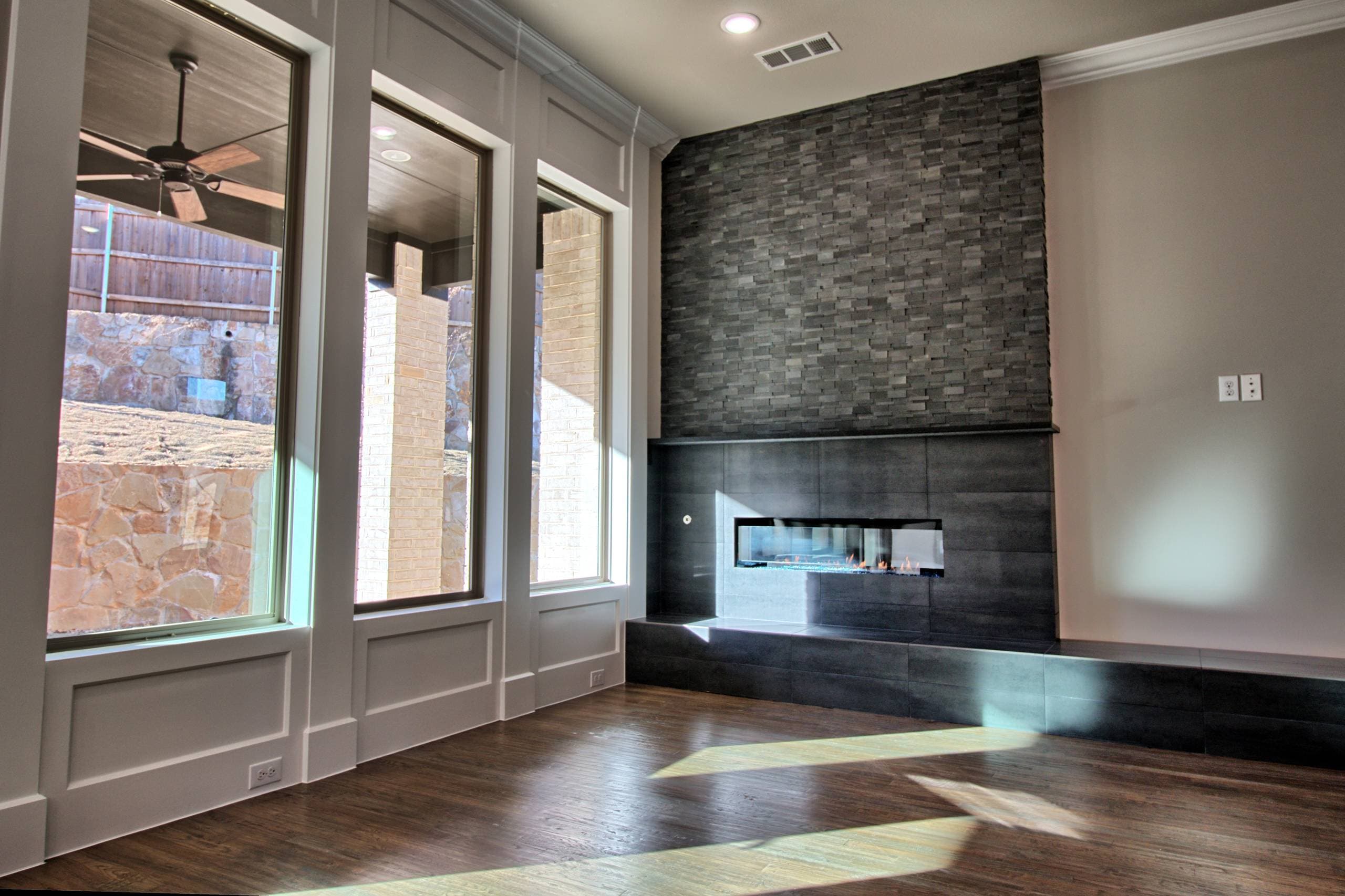 Lit linear gas fireplace in charcoal tile with slim mantel, stacked stone above, raised hearth, and tall windows facing a covered patio