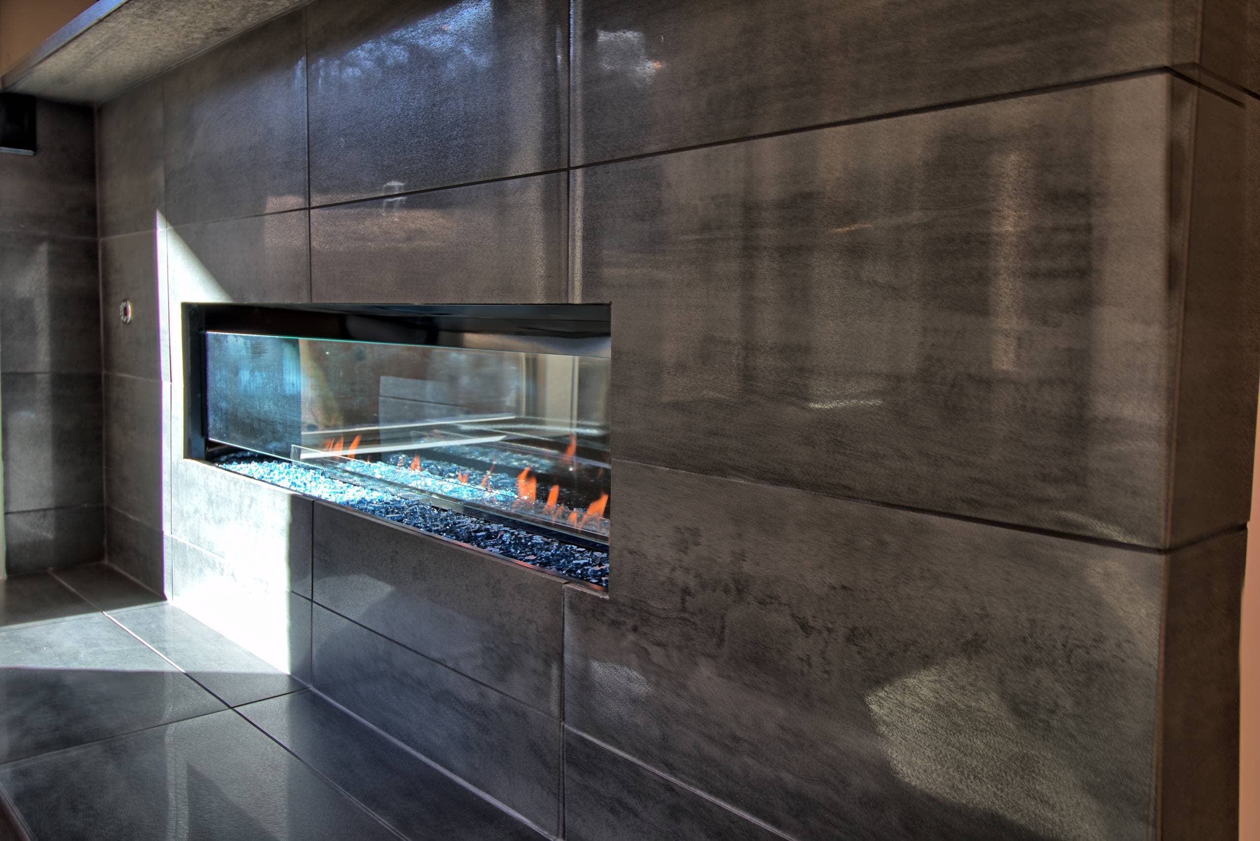 Close-up of a linear gas fireplace with blue fire glass and low flames, framed by large-format charcoal tile with a tiled hearth