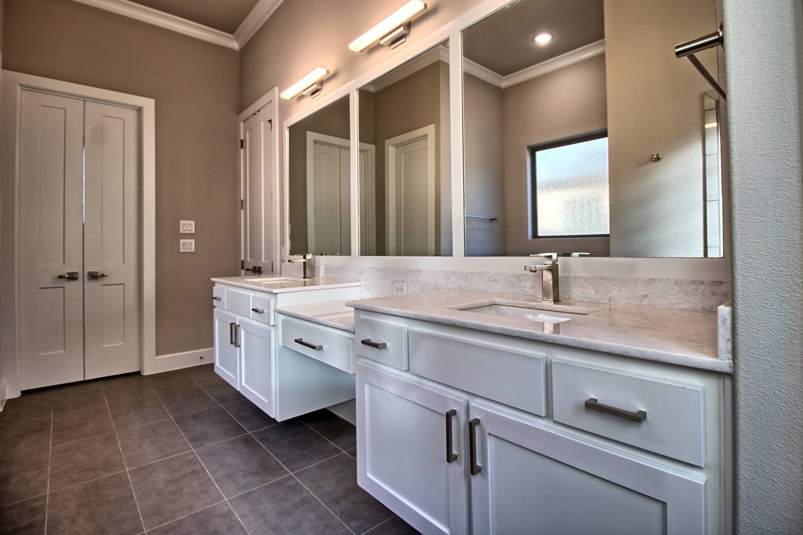Primary bath with white shaker double vanity and makeup space, light stone counters, brushed nickel faucets, linear sconces, and charcoal tile floor