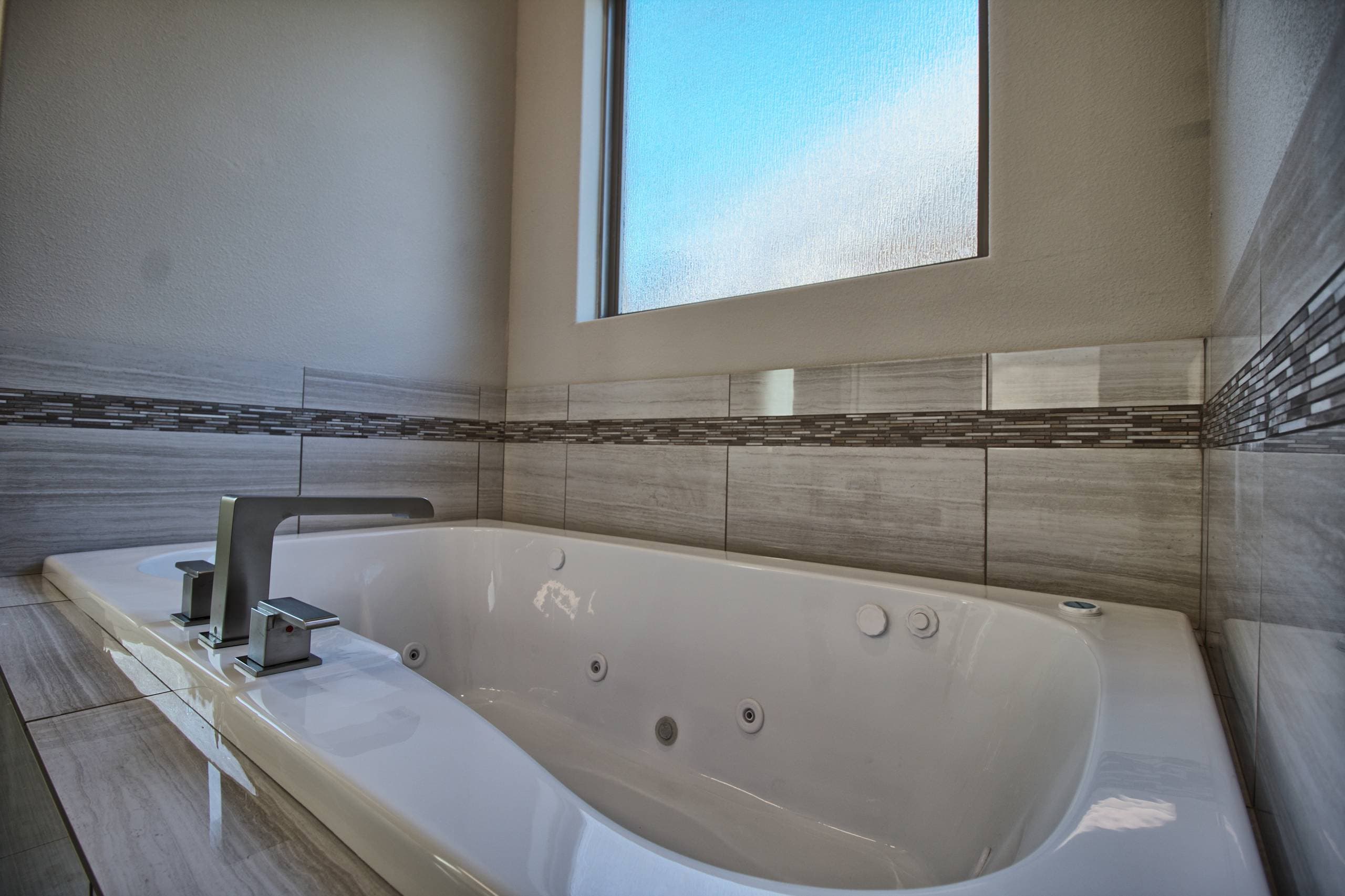 Jetted soaking tub with brushed nickel deck faucet, gray linear-veined tile surround with mosaic accent band, and a frosted glass window above