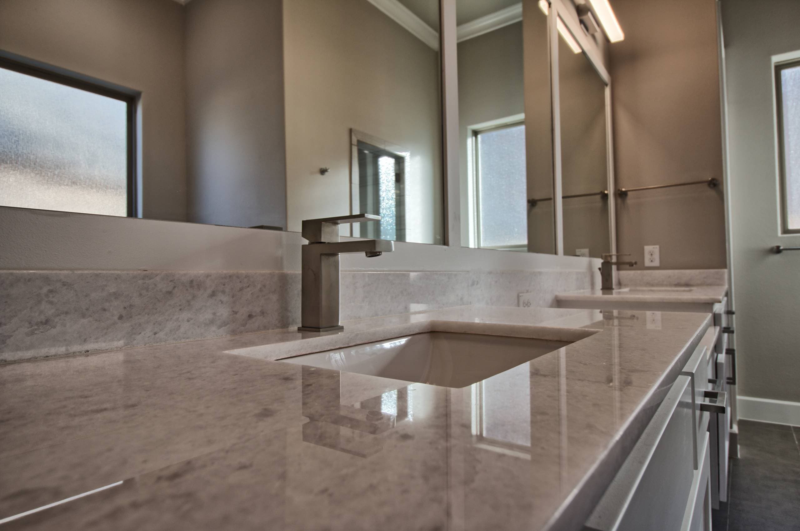 Countertop-level view of a light stone vanity with rectangular undermount sink, brushed nickel single-lever faucet, and wide mirror reflecting frosted windows