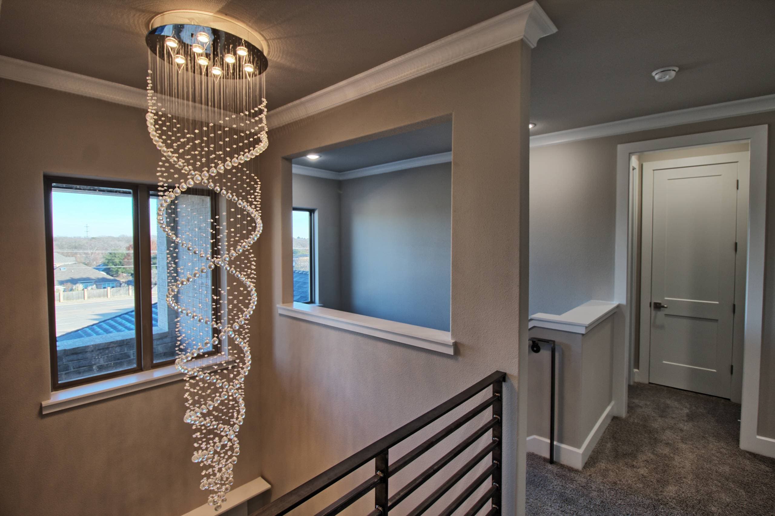 Stairwell with a tall spiral crystal chandelier, horizontal dark metal railing, and a window, beside a carpeted upstairs hallway