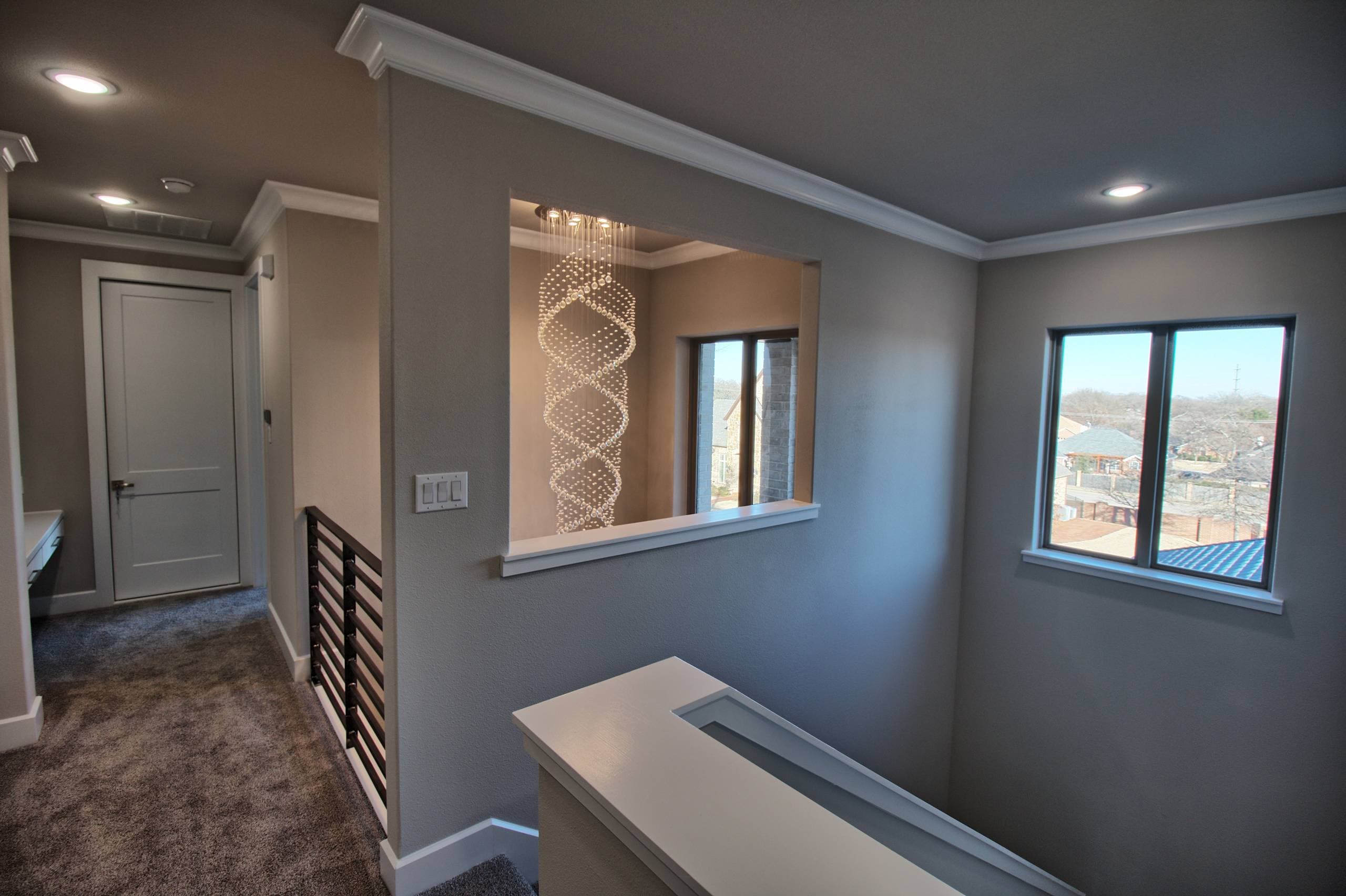 Carpeted upstairs landing with an interior wall opening framing a spiral crystal chandelier, horizontal metal stair railing, and a window