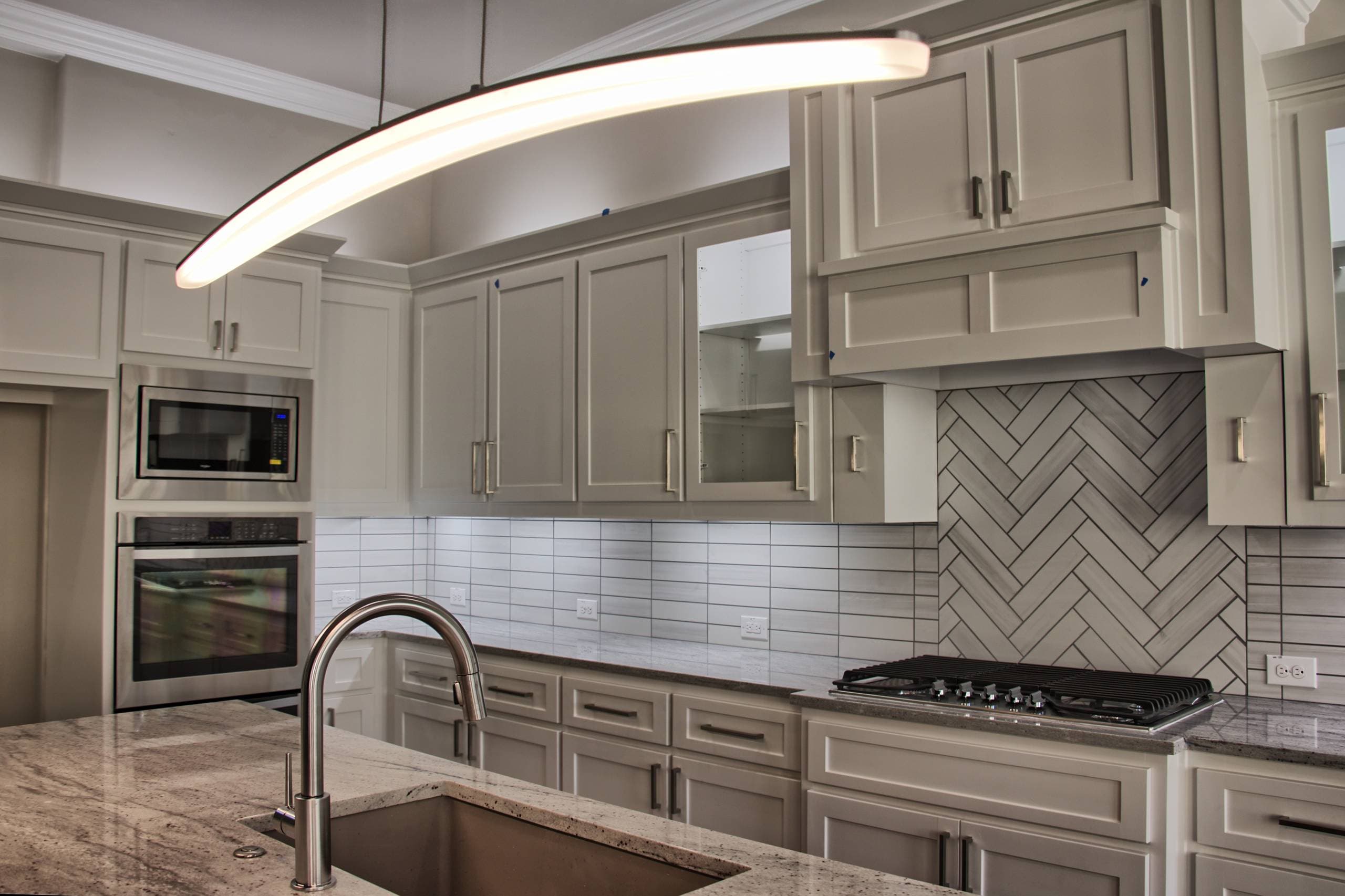 Curved LED pendant over a granite island sink, facing light gray shaker cabinets, herringbone tile behind a gas cooktop, and stainless appliances
